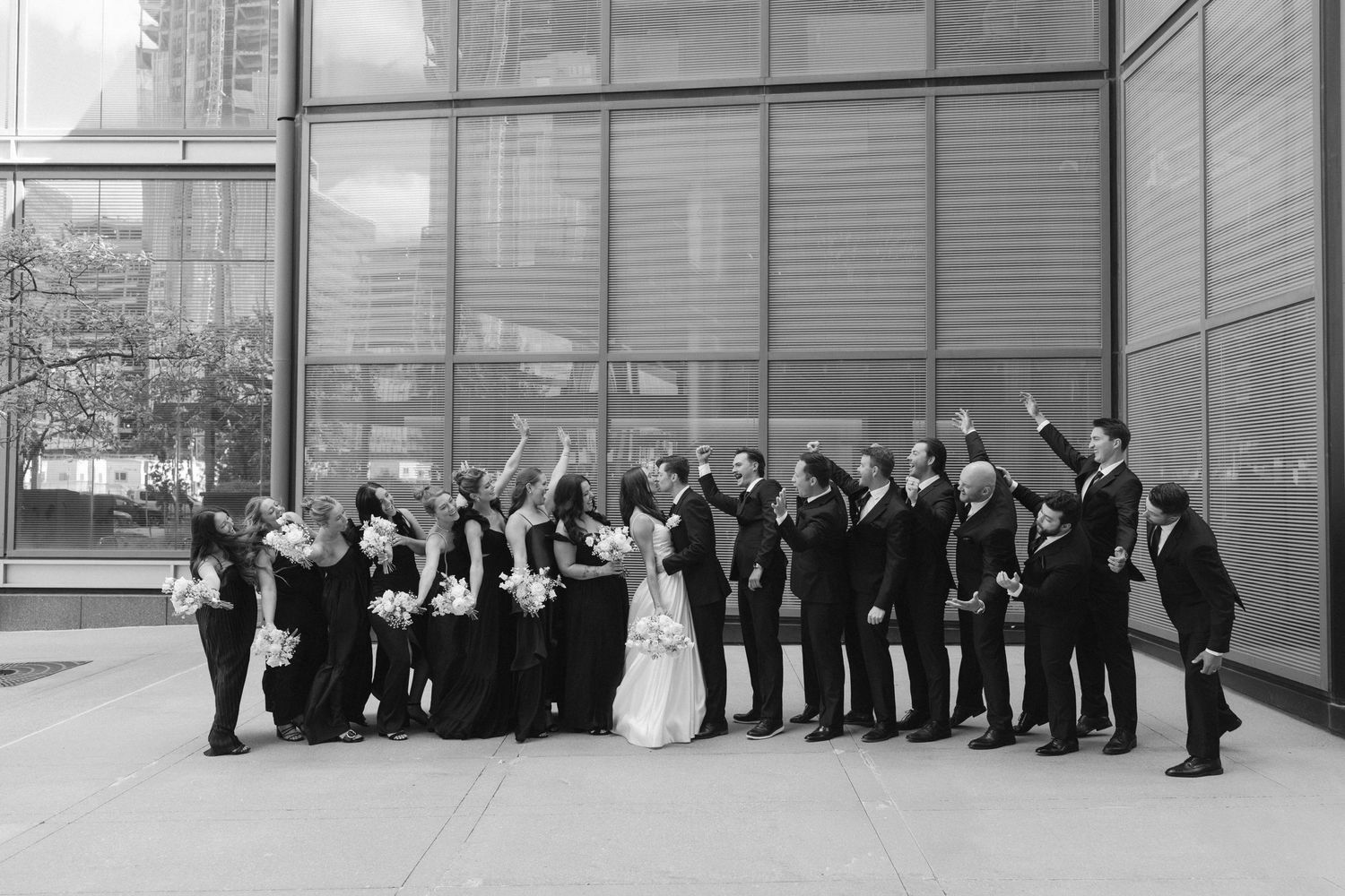 Wedding party lined up against modern glass building exterior for formal group photos.