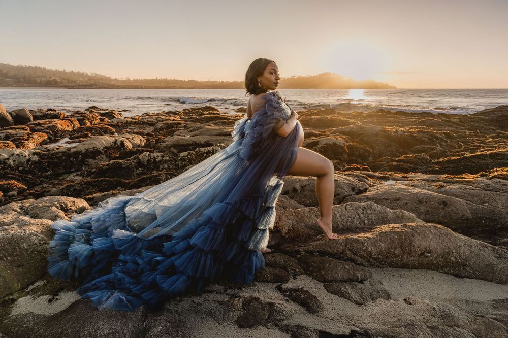 Pregnant woman in flowing blue tulle gown poses on rocky coastal shore at golden sunset.