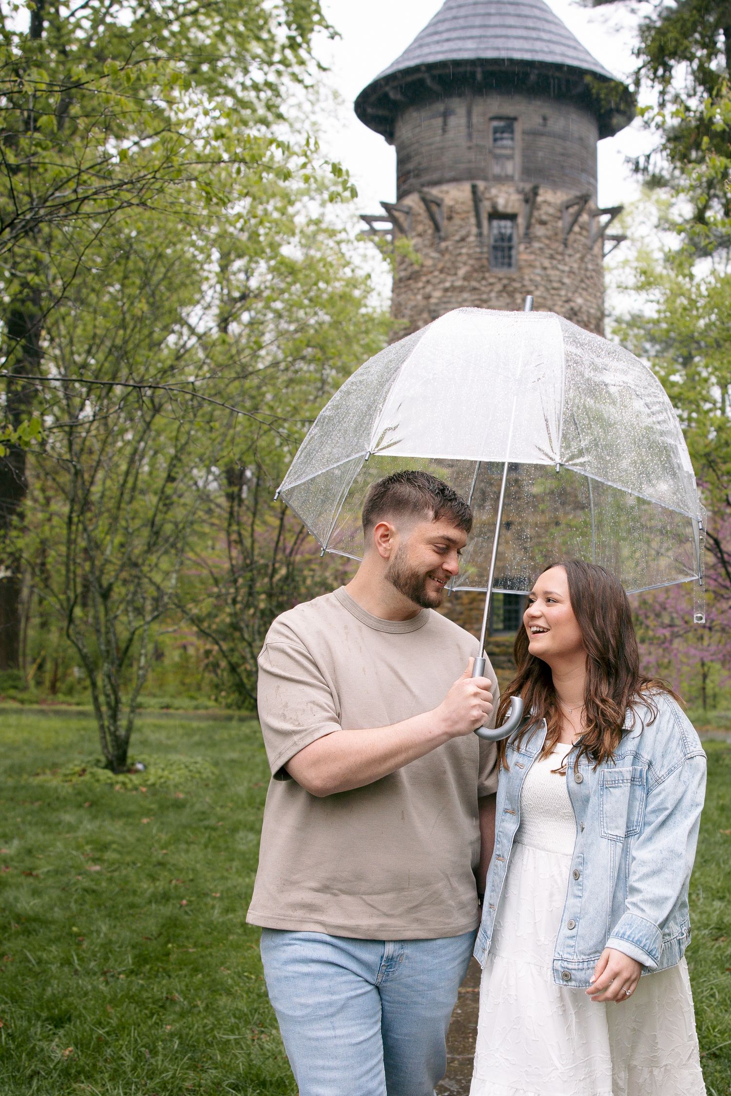 Happy couple sharing a clear umbrella in a lush green park with a historic stone tower in the background.