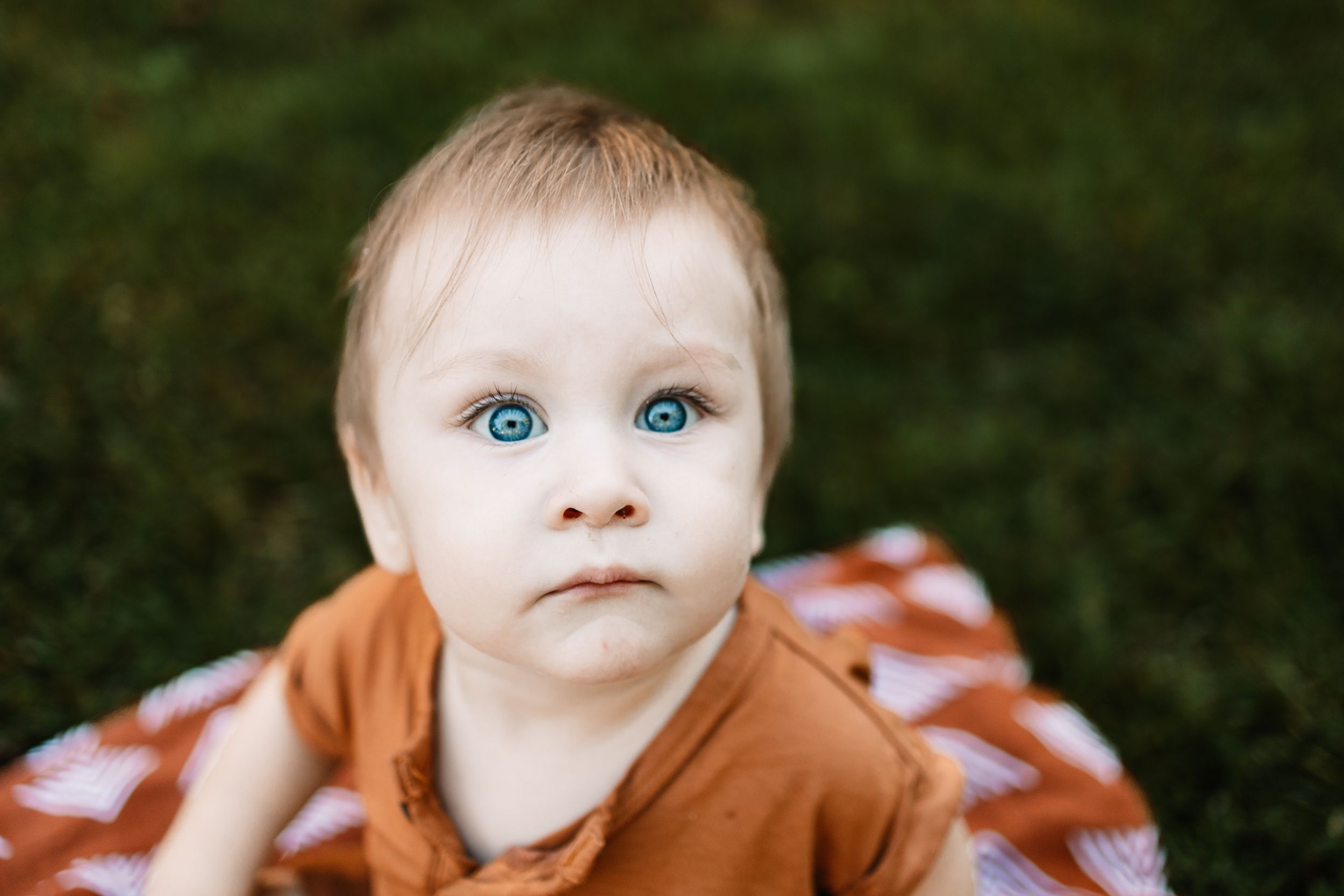 Adorable baby with striking blue eyes wearing an orange outfit, lying on a colorful blanket on green grass.