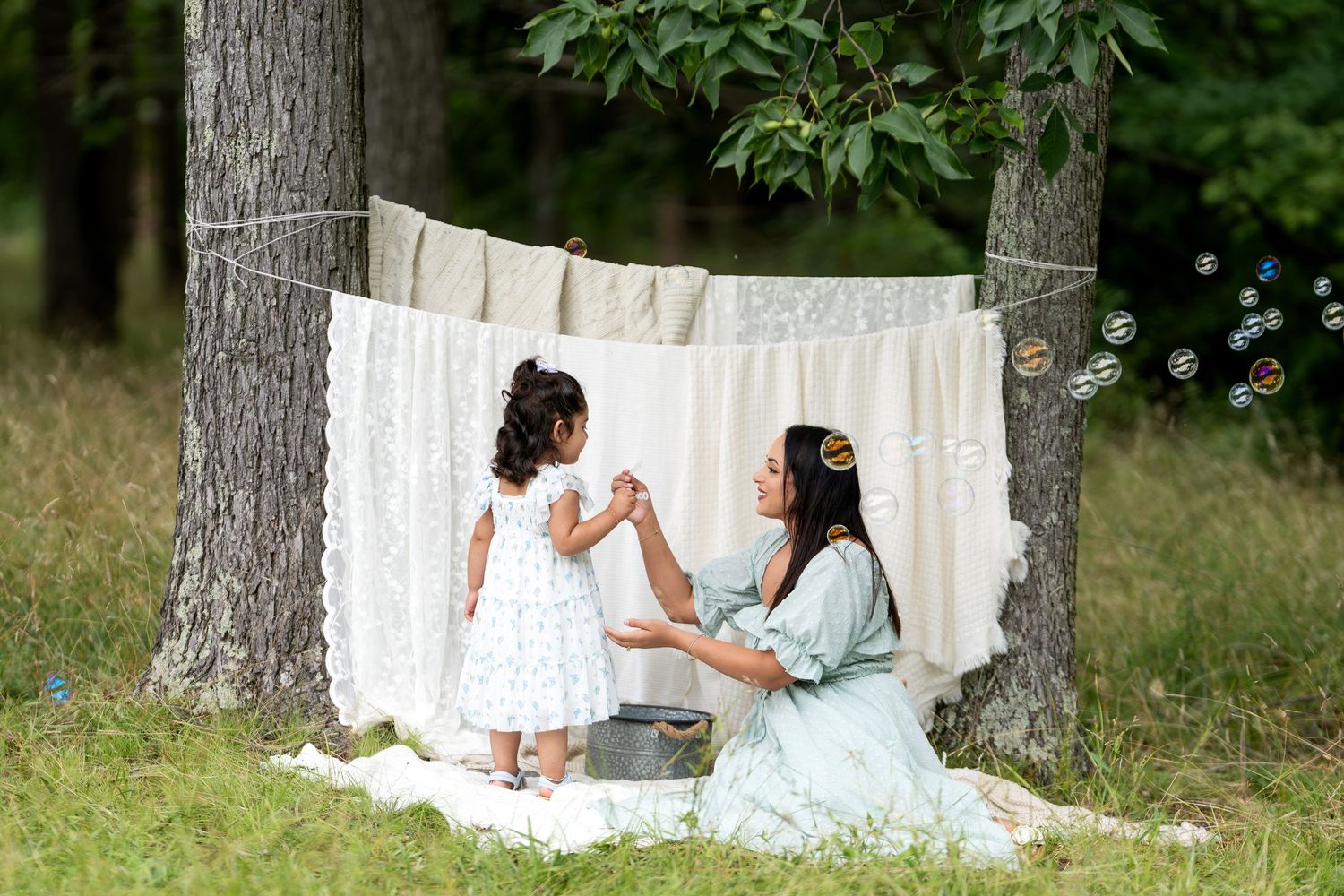Mother and daughter playing with bubbles outdoors near lace fabric hung between trees in a forest setting.