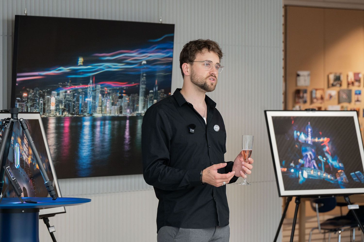 Man in black shirt holding champagne glass speaks at art gallery with vibrant city skyline photograph displayed behind him.