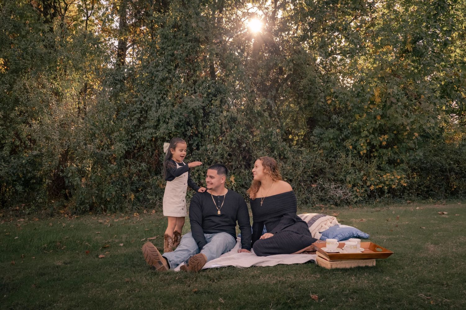 Family enjoying a golden hour picnic on grass, with children playing and laughing surrounded by lush green trees. Morgantown, WV
