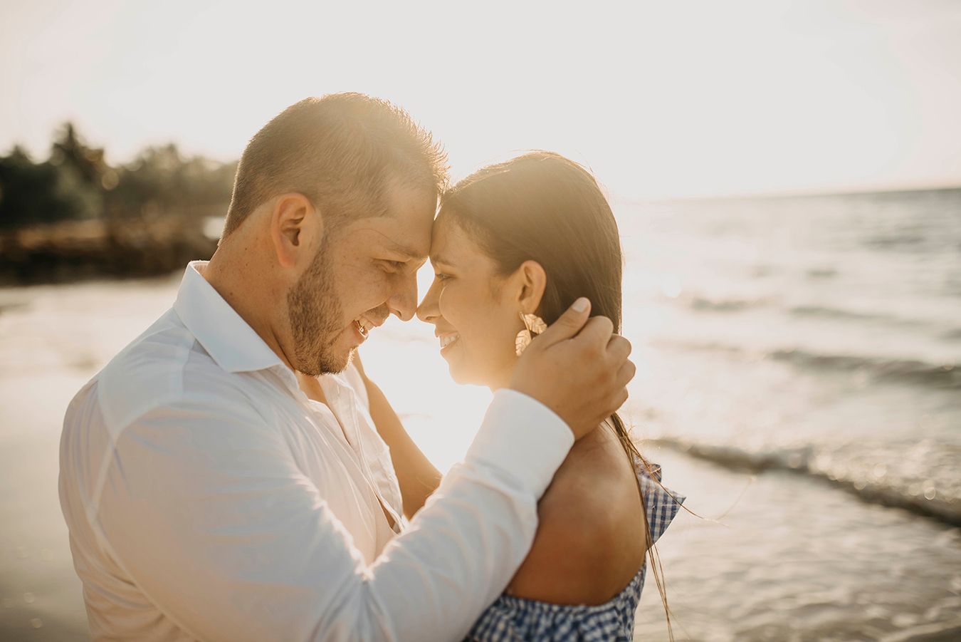 Romantic couple embracing forehead-to-forehead on a sunlit beach at golden hour near the ocean.