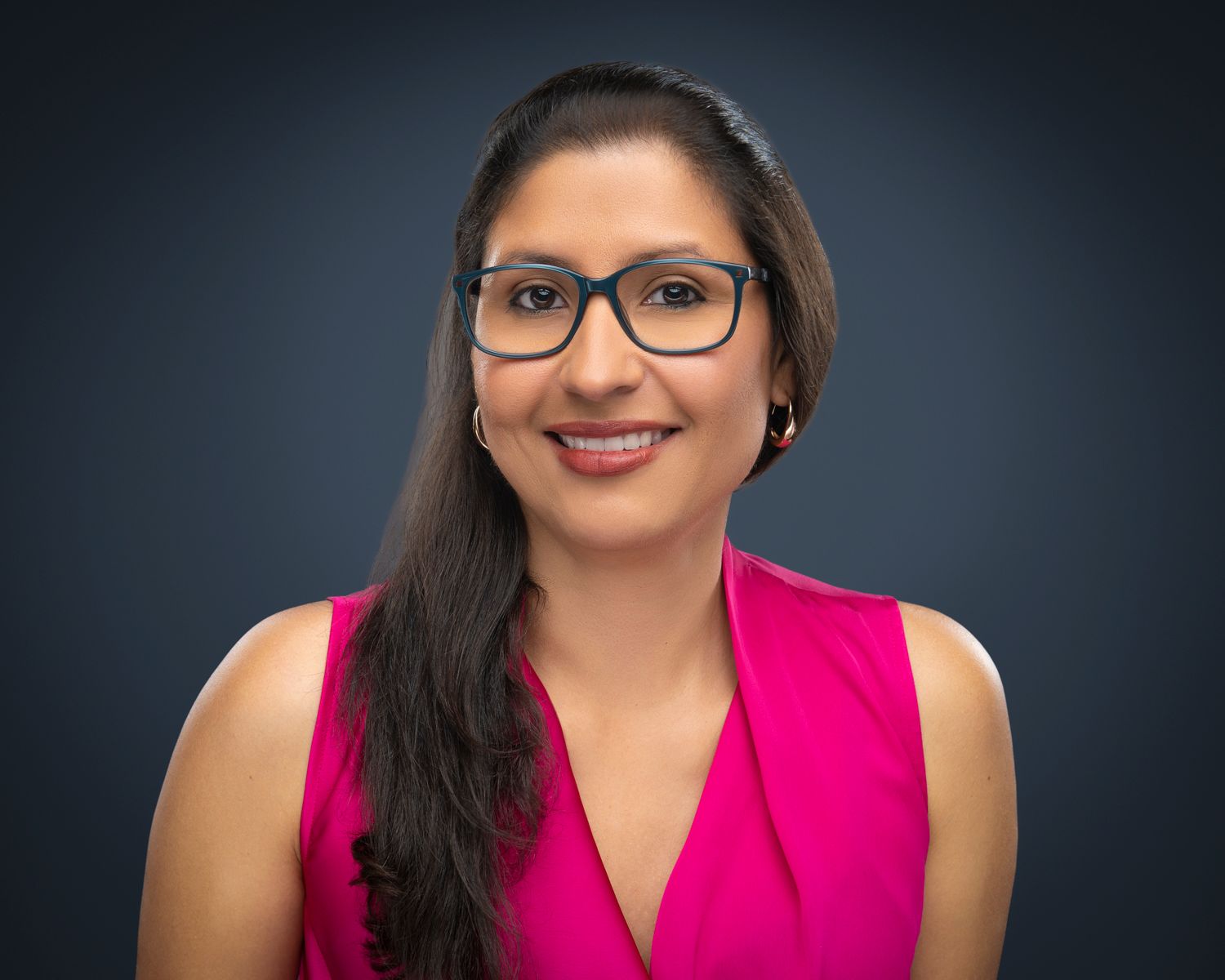 Professional headshot of a smiling woman with glasses and long dark hair wearing a bright pink sleeveless top.