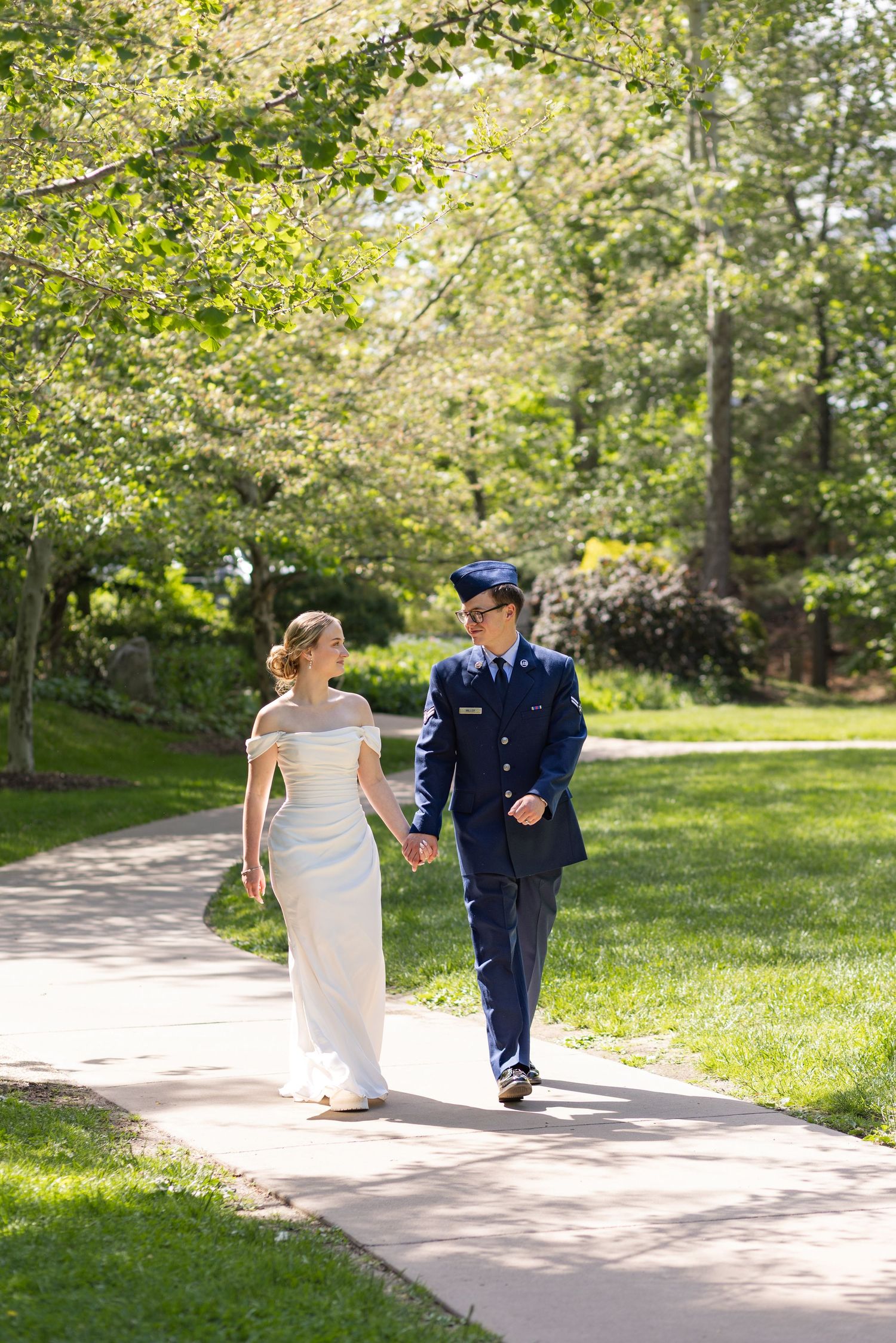 Newlywed couple walking hand in hand on a sunny park path, bride in white gown and groom in military dress uniform.