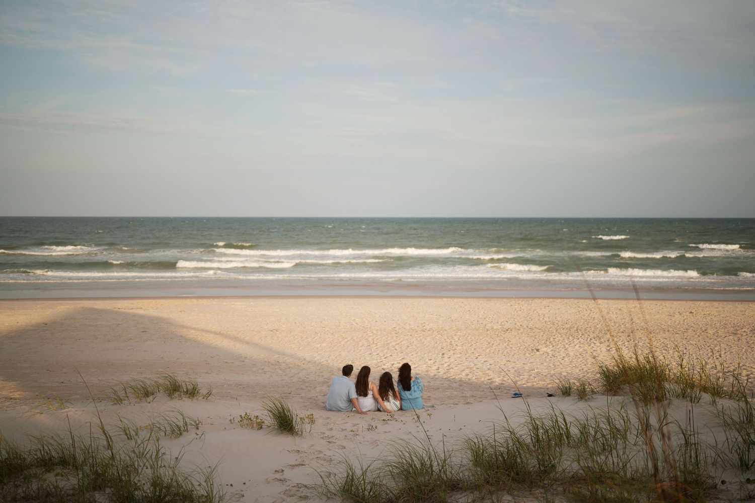 Family of four sitting together facing the ocean on a serene sandy beach at dusk.