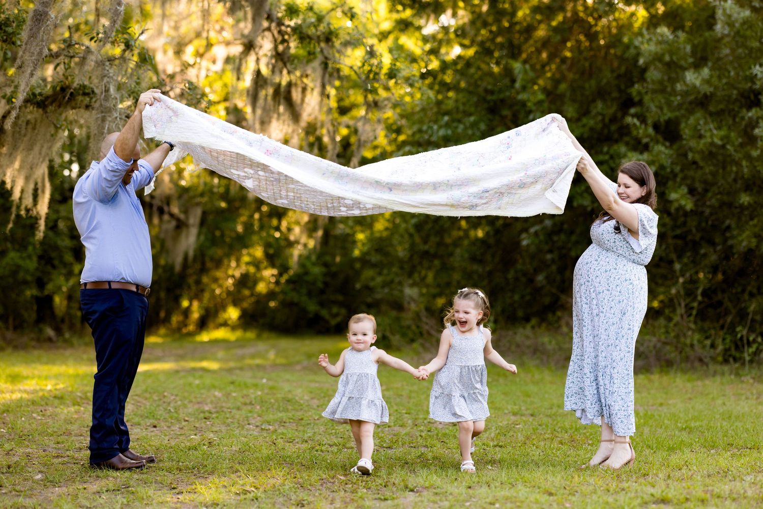 Family of four with pregnant mom holding a blanket over two young girls in a sunny green park.