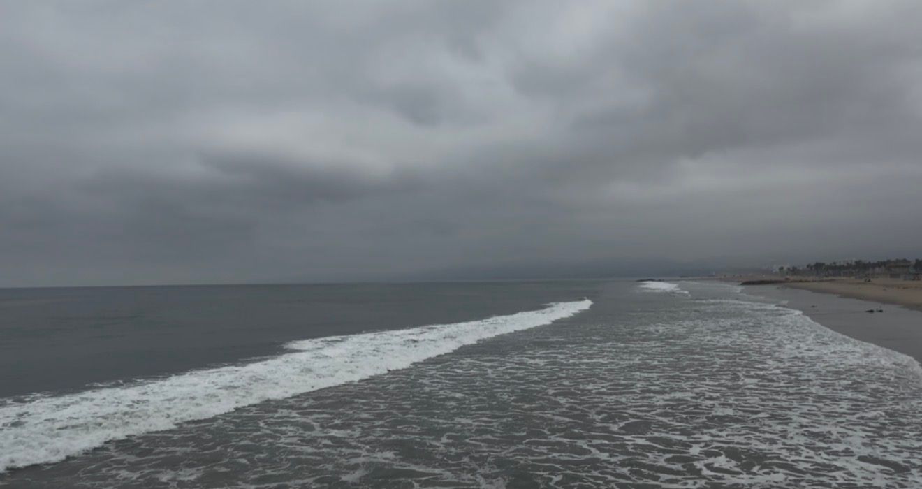 Aerial view of overcast beach with gentle waves washing ashore under dramatic gray cloudy skies.