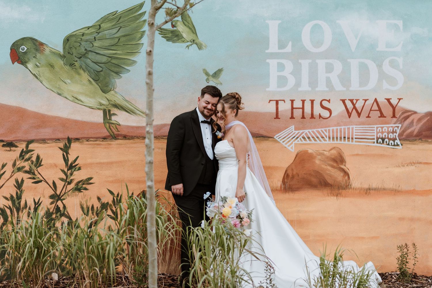 Bride and groom pose in front of a colorful Love Birds mural with desert landscape at their wedding.