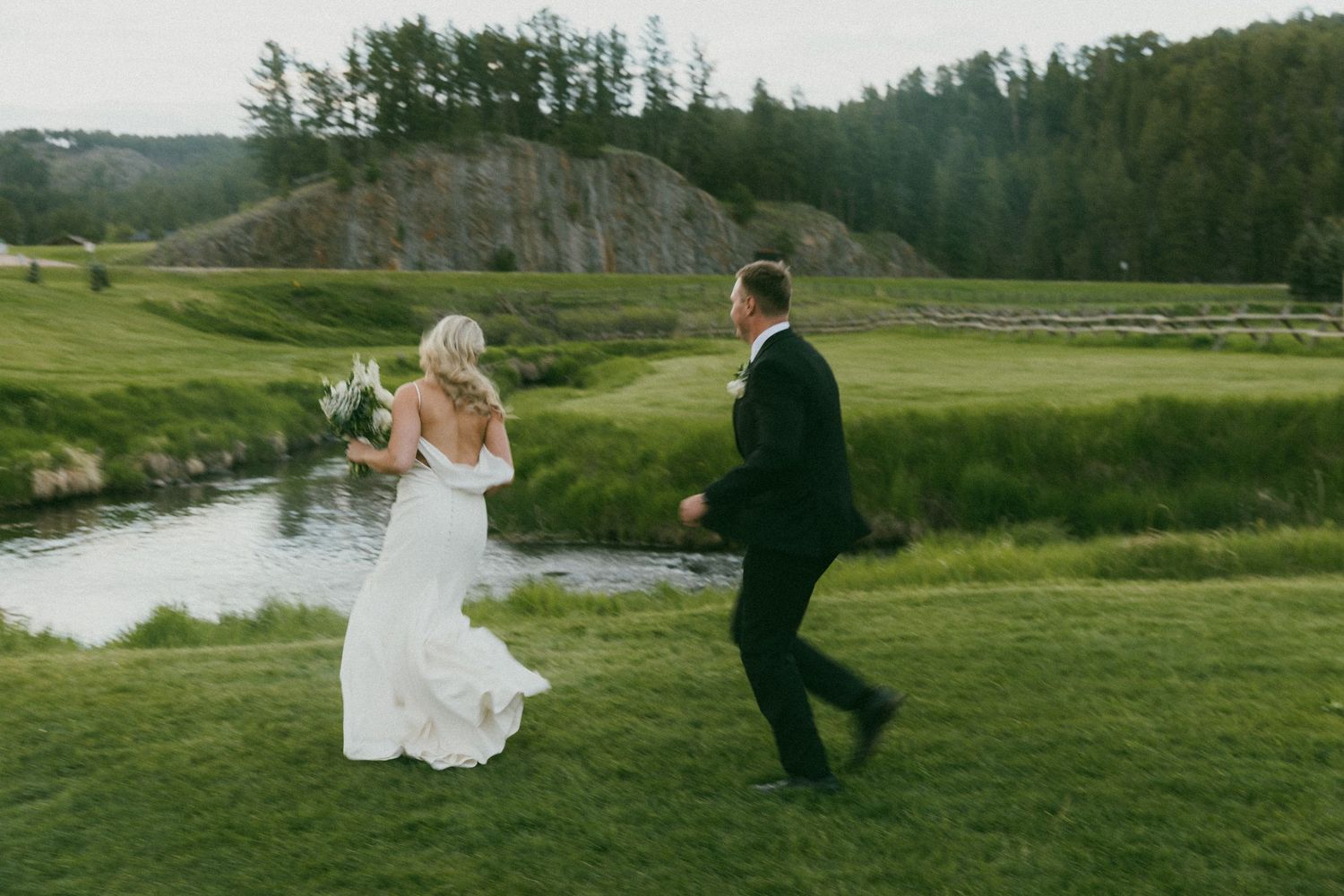 Couple running through a lush green meadow with mountains in the background.