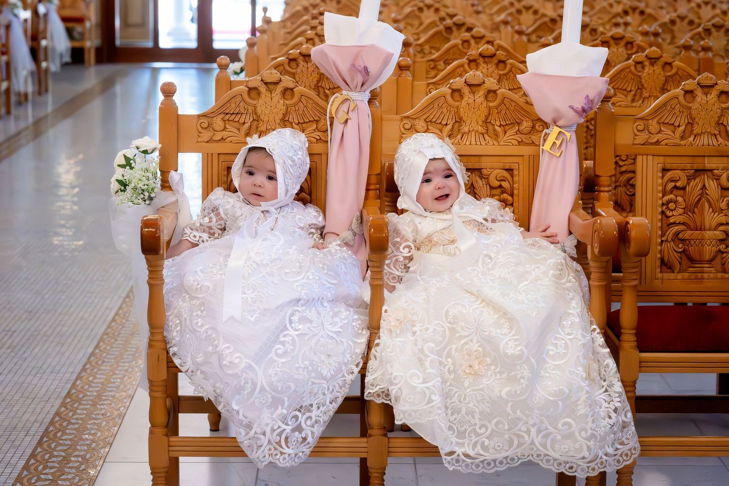 Two baby girls in matching white lace christening gowns and bonnets seated on wooden church pews during a baptism ceremony.