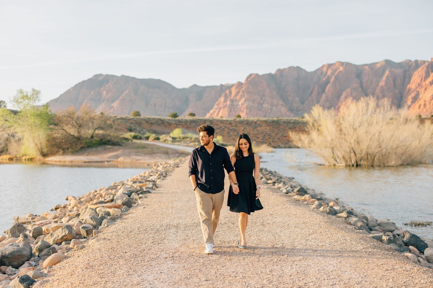 Couple in matching black outfits walking hand-in-hand on a rocky path by a lake with red mountains behind.