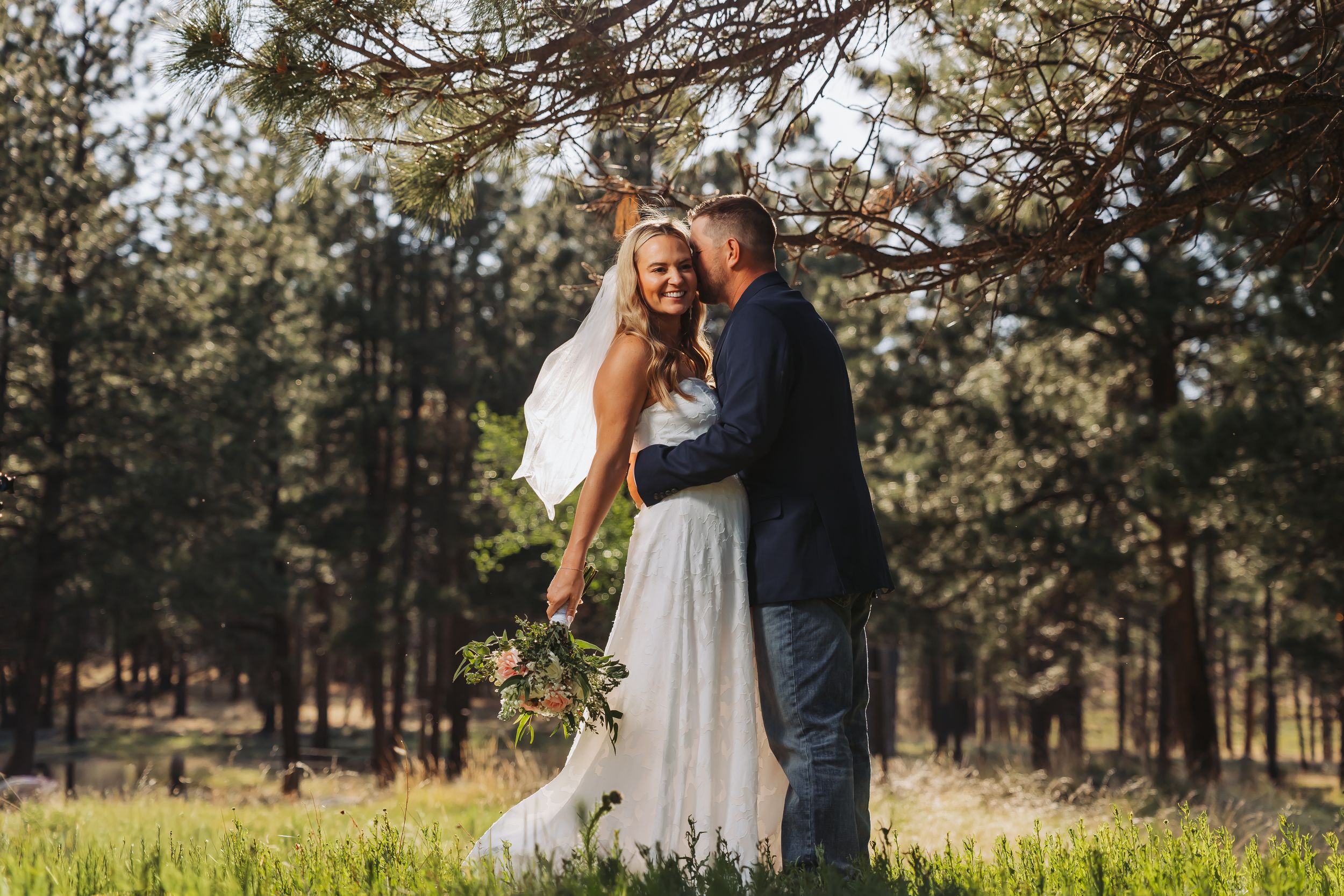 A bride and groom embrace in a sunlit forest meadow, the bride holding a bouquet in her white gown.