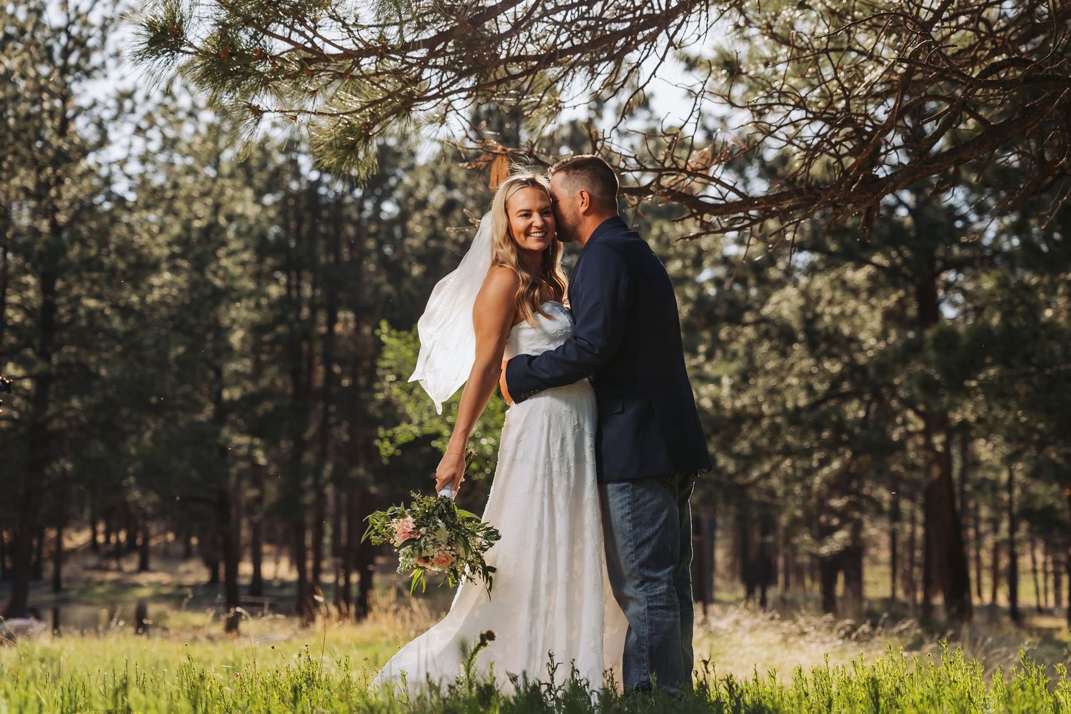 A bride and groom embrace in a sunlit forest meadow, the bride holding a bouquet in her white gown.