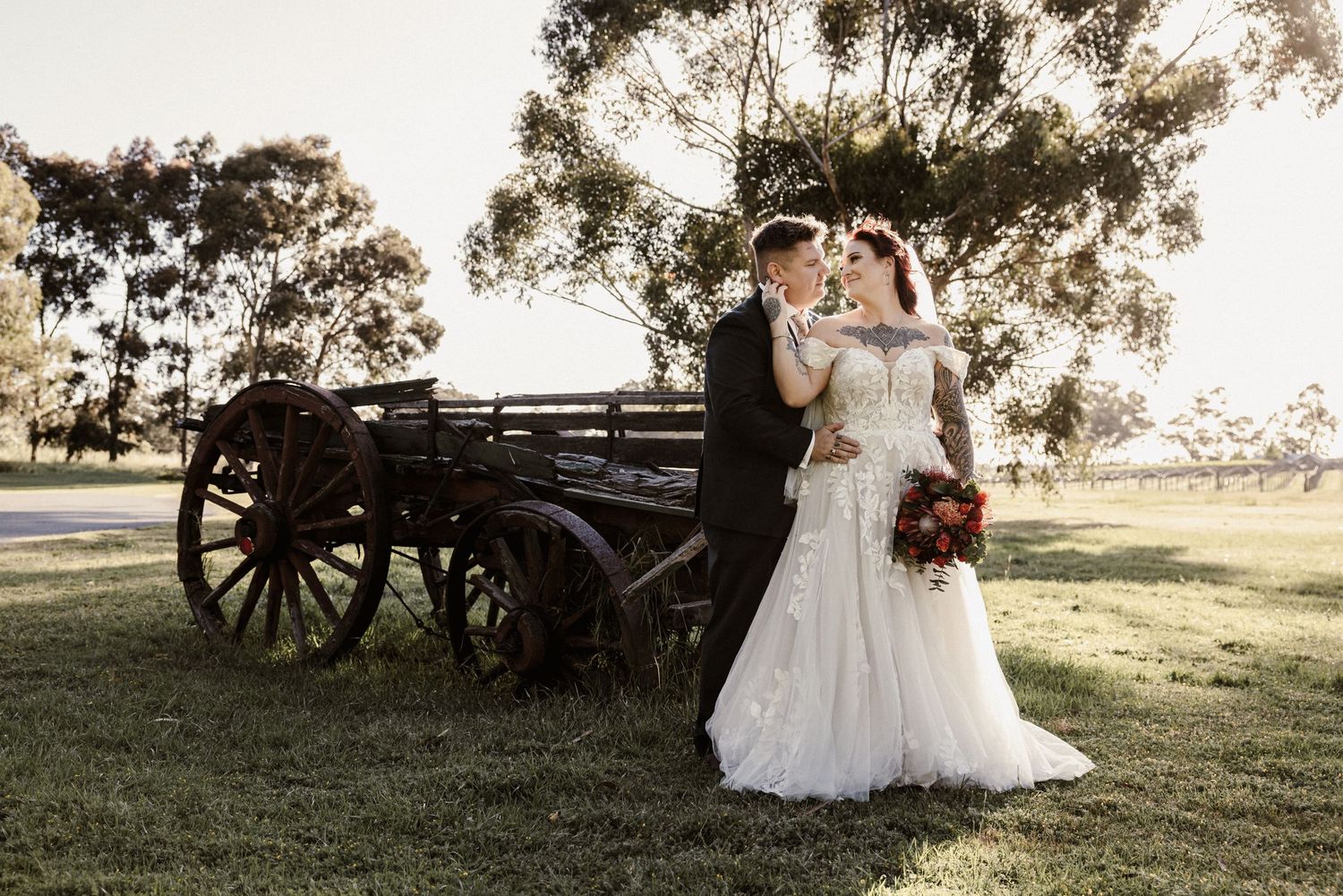 A bride and groom share a romantic kiss surrounded by lush greenery with a vibrant red and orange bridal bouquet.