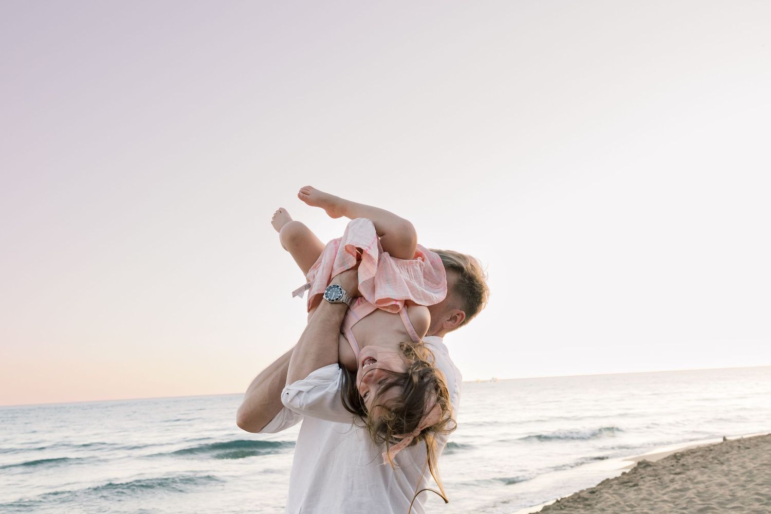 A joyful parent lifts a laughing toddler overhead on a serene beach at sunset.