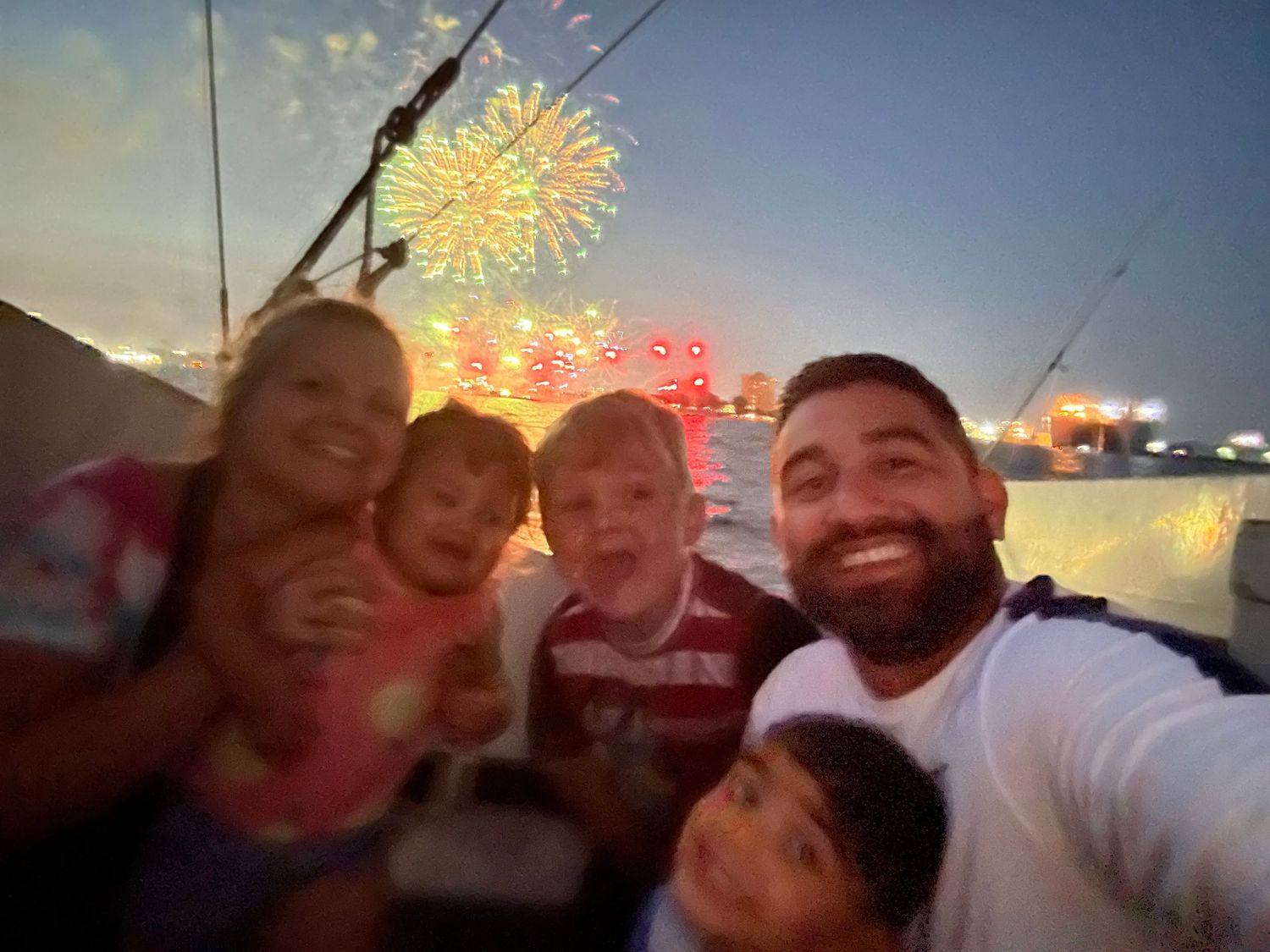 Family selfie with fireworks lighting up the evening sky in the background, celebrating outdoors together.