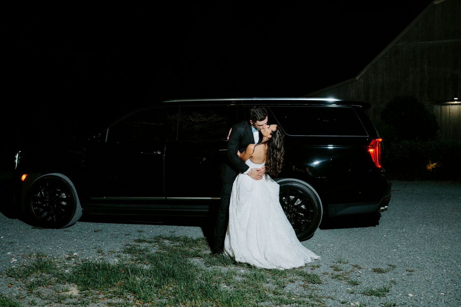 Bride and groom share a romantic kiss in front of a black SUV limousine at night during their wedding.