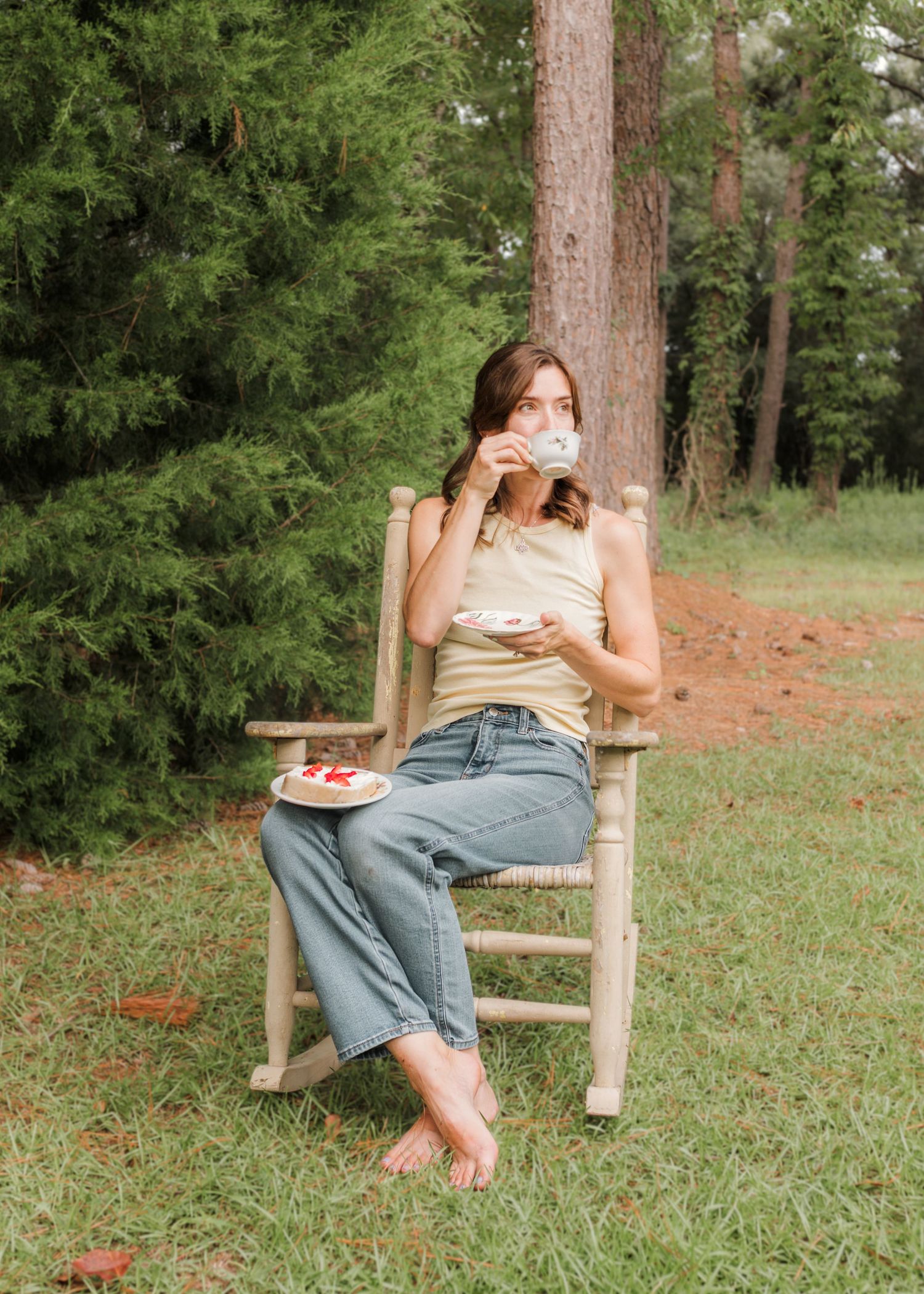 Woman sitting barefoot in wooden rocking chair outdoors, drinking from cup with plate of food, surrounded by green trees.