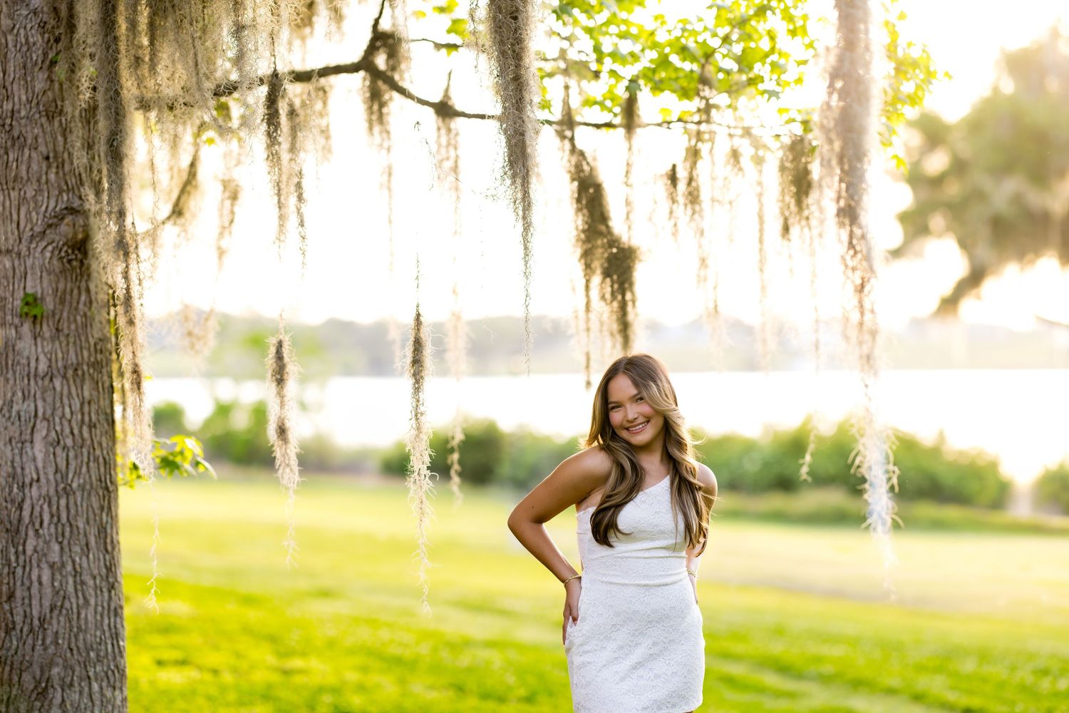 Young woman in white dress posing under Spanish moss-draped tree with sunlit green lawn and lake backdrop.