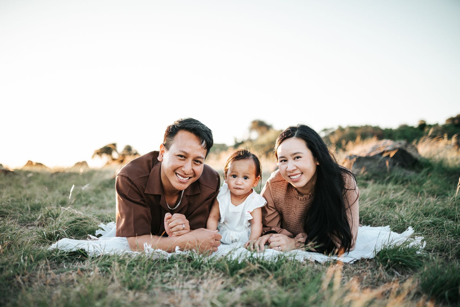 Happy Melbourne family with baby lying on blanket in green field at sunset, smiling outdoors taken in Altona by Melbourne photographer.