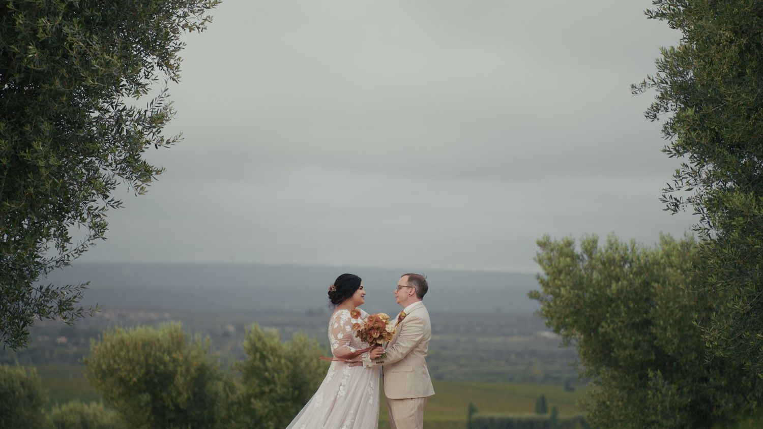 A couple in wedding attire holds hands outdoors, surrounded by trees with a scenic countryside landscape in the background.