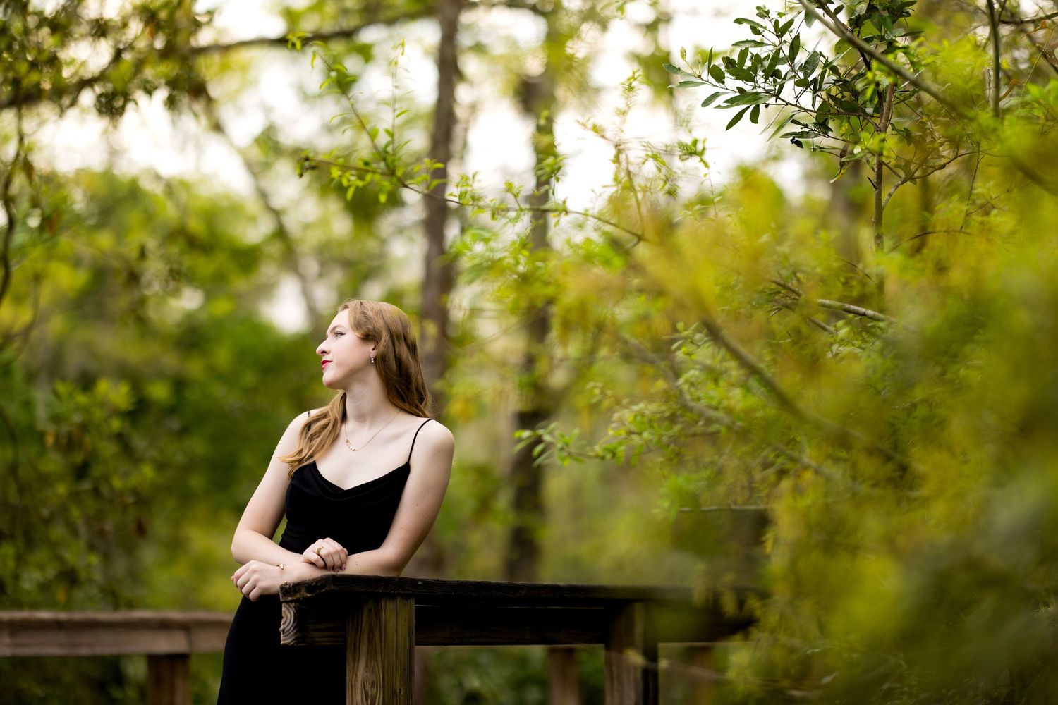 A person in a black dress poses outdoors against a green natural backdrop in a romantic photoshoot setting.