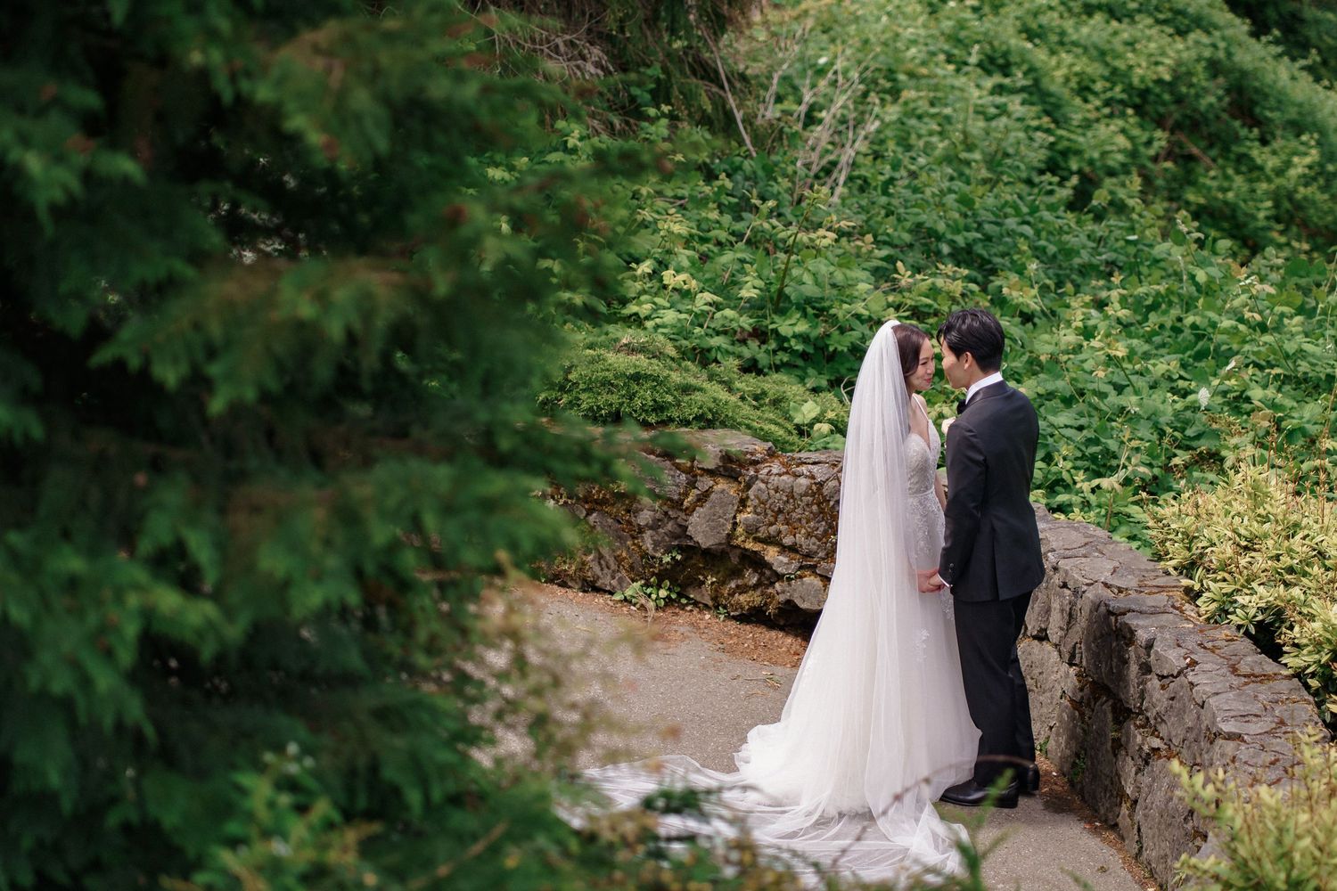A couple in wedding attire stands together in a lush green forest setting near a stream.Queen Elizabeth Park Wedding