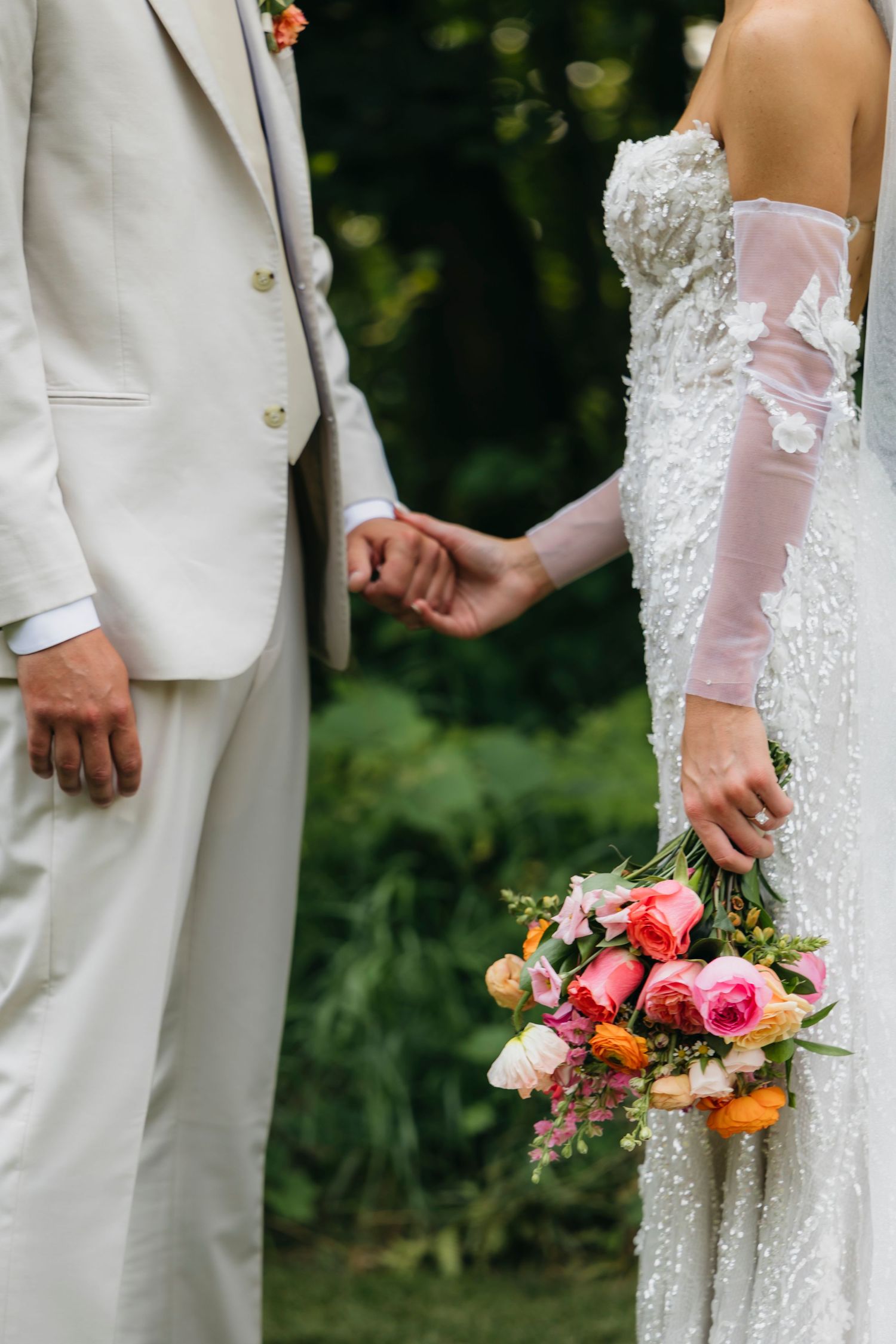 Wedding party members in white attire stand in line holding vibrant coral and orange floral bouquets.