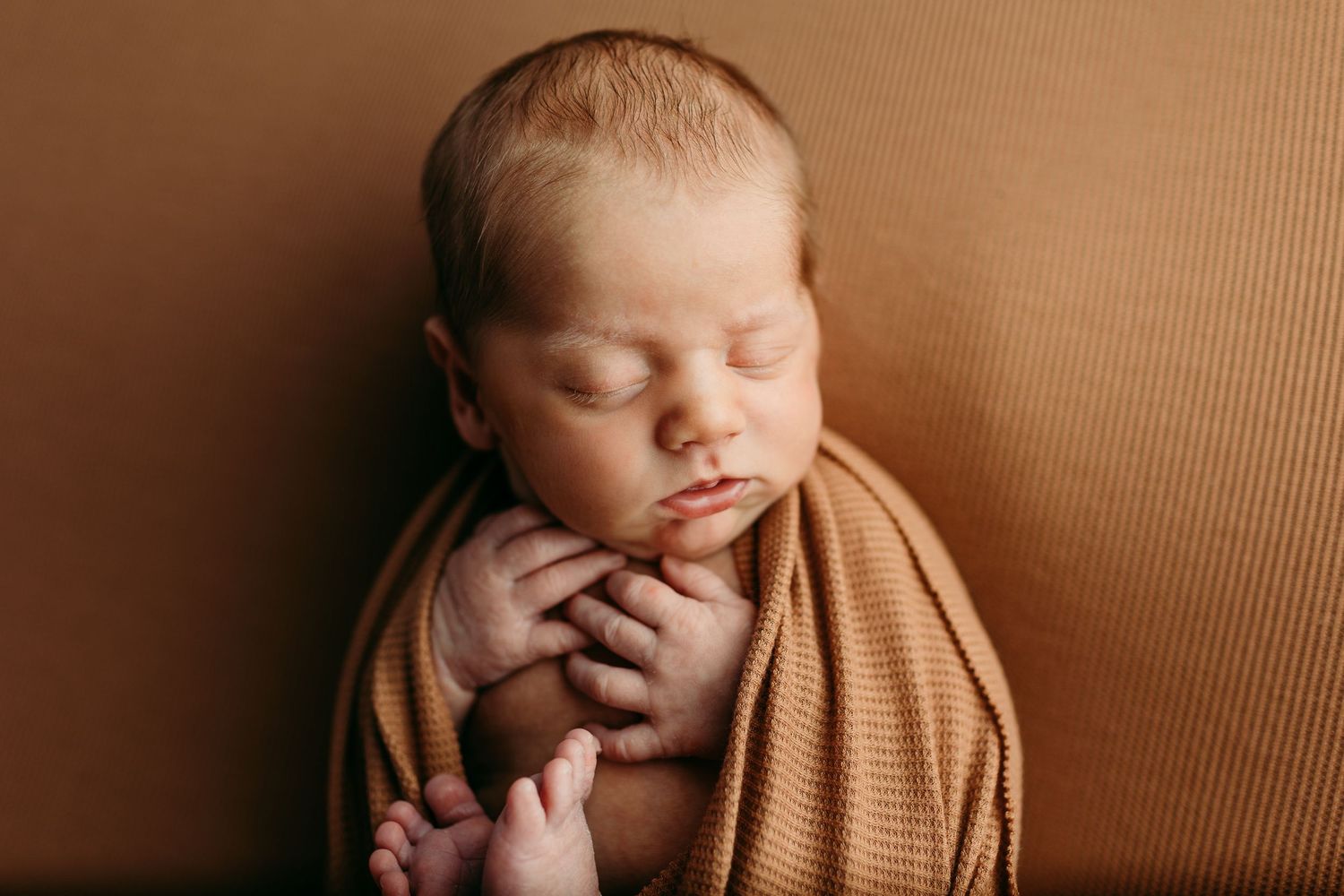 Peaceful newborn baby wrapped in a warm tan swaddle blanket, sleeping with tiny hands tucked under chin.