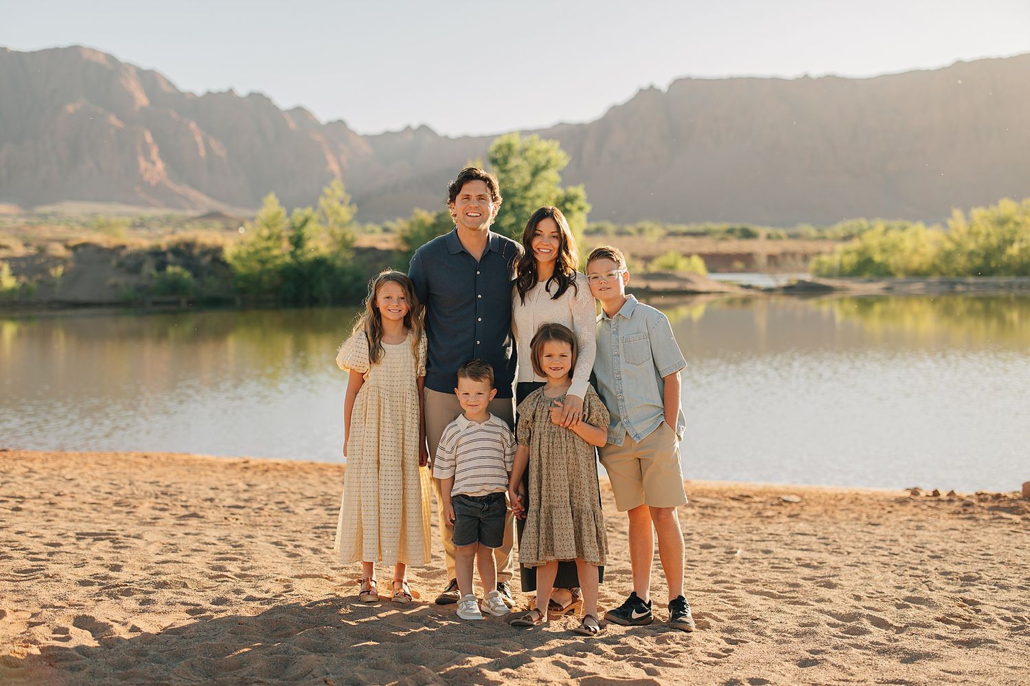 Family of six posing on sandy beach near river with mountain backdrop in warm golden light.