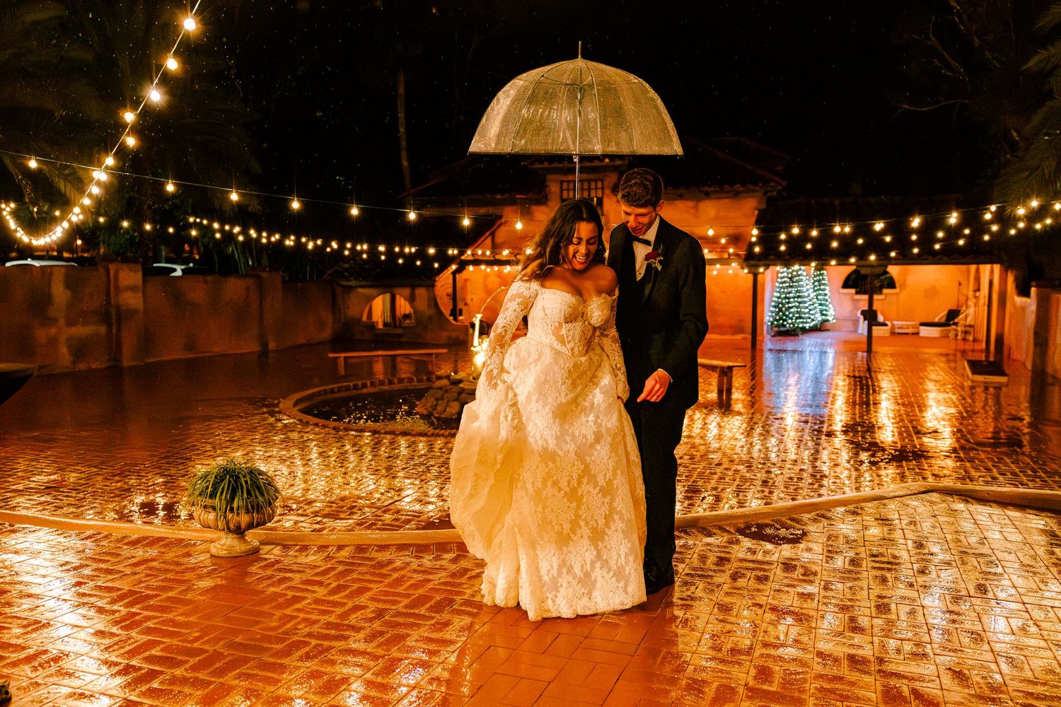 Bride and groom walking with clear bubble umbrella through rain, with string lights above them, lots of ambient light at night