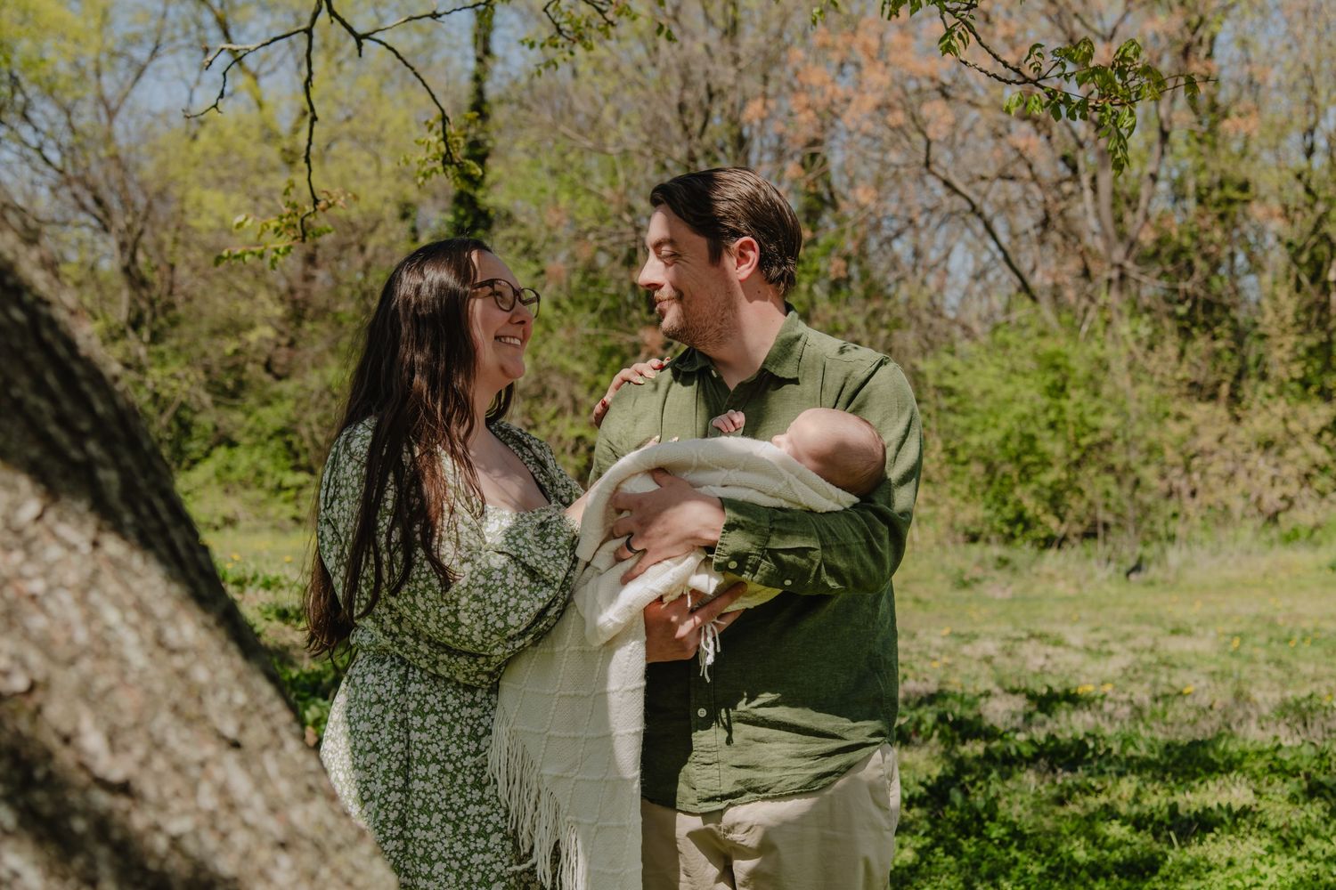 A couple smiles at each other while holding a newborn baby outdoors in a lush green park setting.