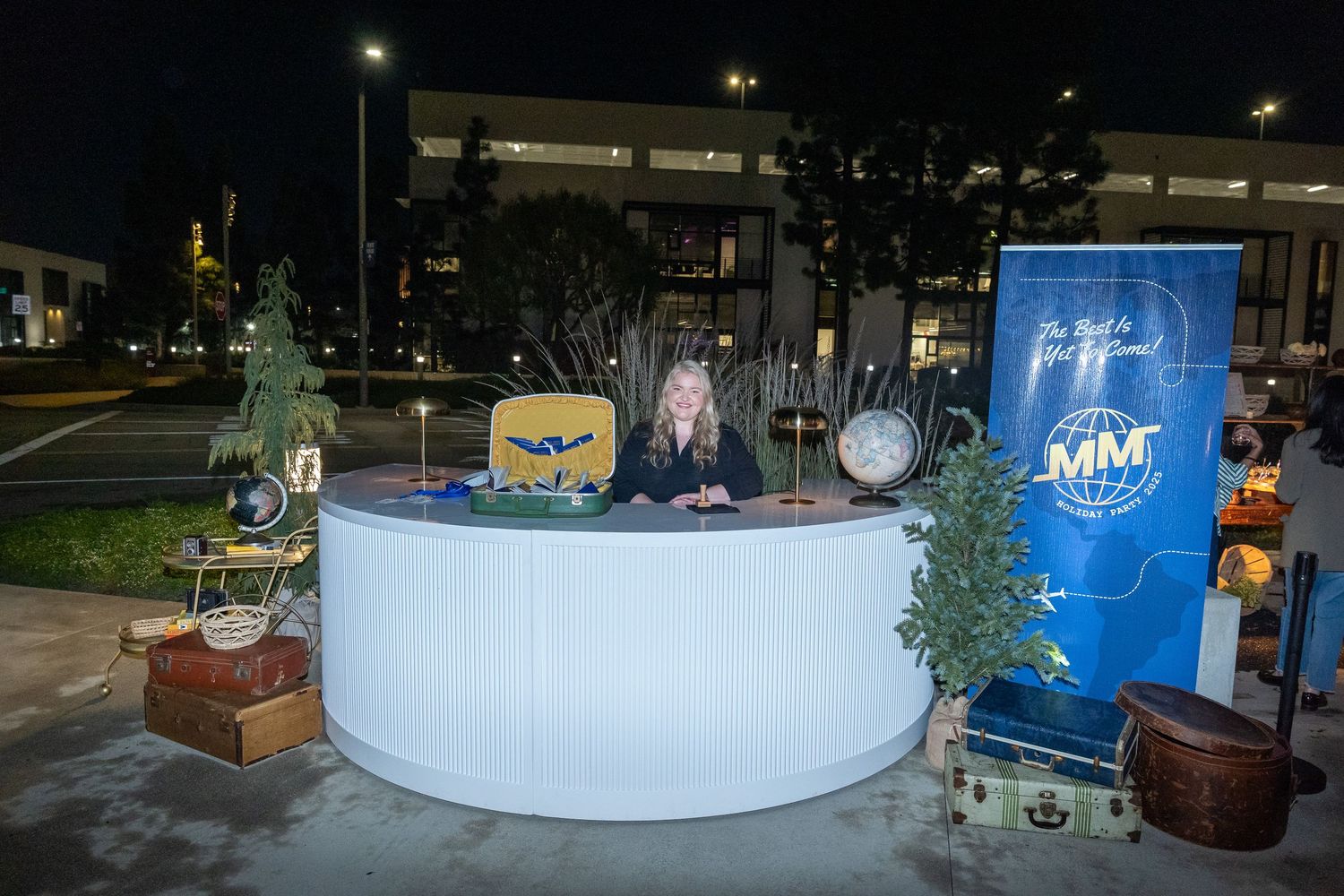 Woman at a circular white reception desk outdoors at night with travel-themed decor and MM Global branding banner.