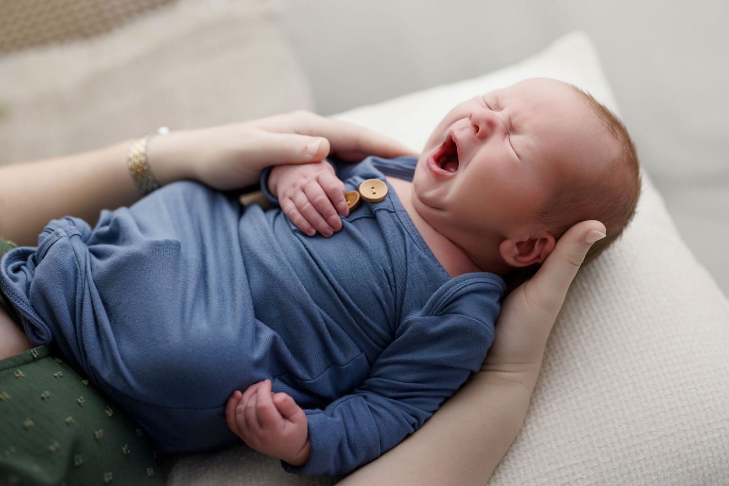 Newborn baby yawning in blue swaddle outfit, cradled in adult arms on soft cushion.