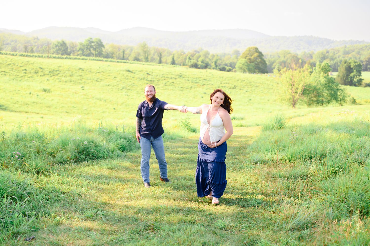 Couple holding hands and dancing playfully in a lush green meadow with mountain views.