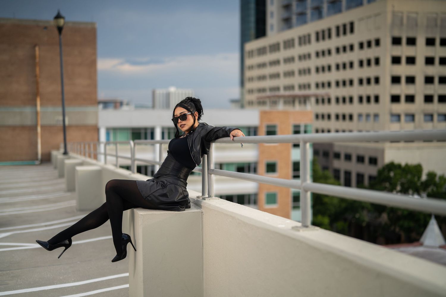Woman in black outfit and heels poses on rooftop ledge with city buildings in background.