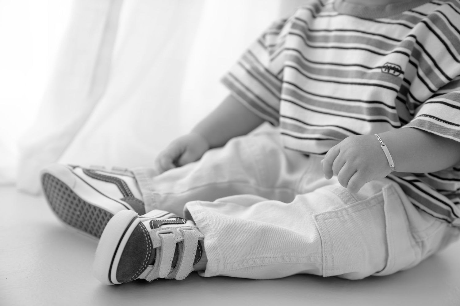 Black and white photo of a young toddler sitting down wearing striped shirt, light pants, and sneakers.