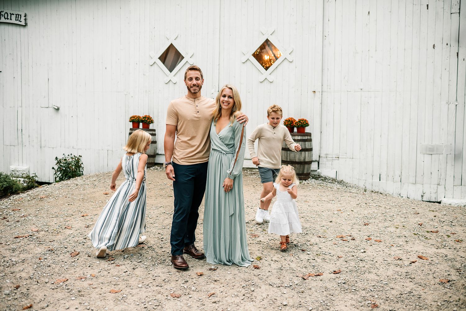 Family of five posing together in front of a white barn, parents with three young children in elegant attire.