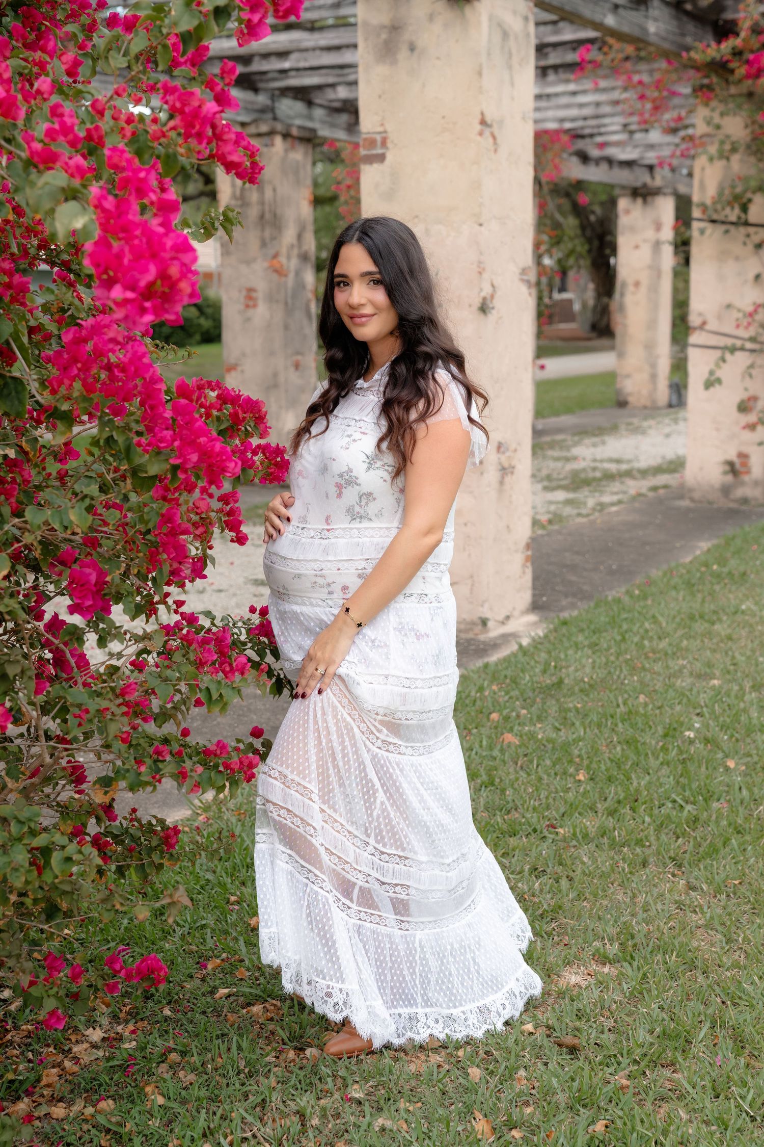 Pregnant woman in white lace dress poses beside vibrant pink bougainvillea flowers in a garden setting.
