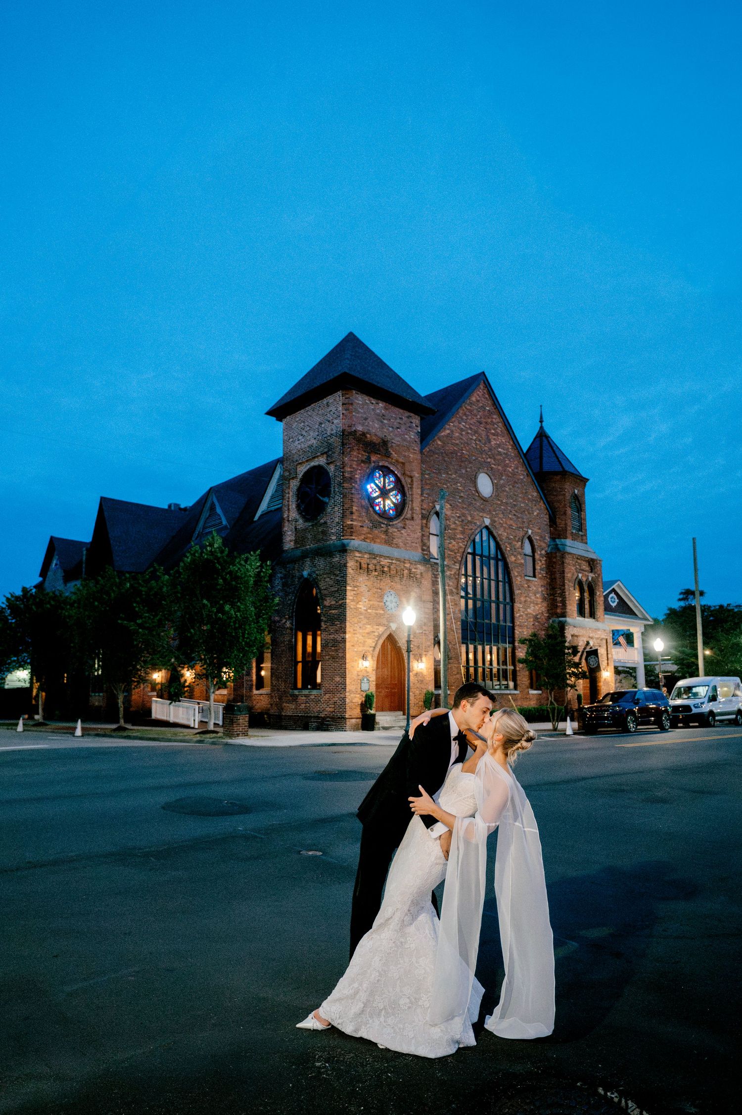 Bride and groom share a romantic kiss outside a historic brick church venue at twilight during their wedding night.