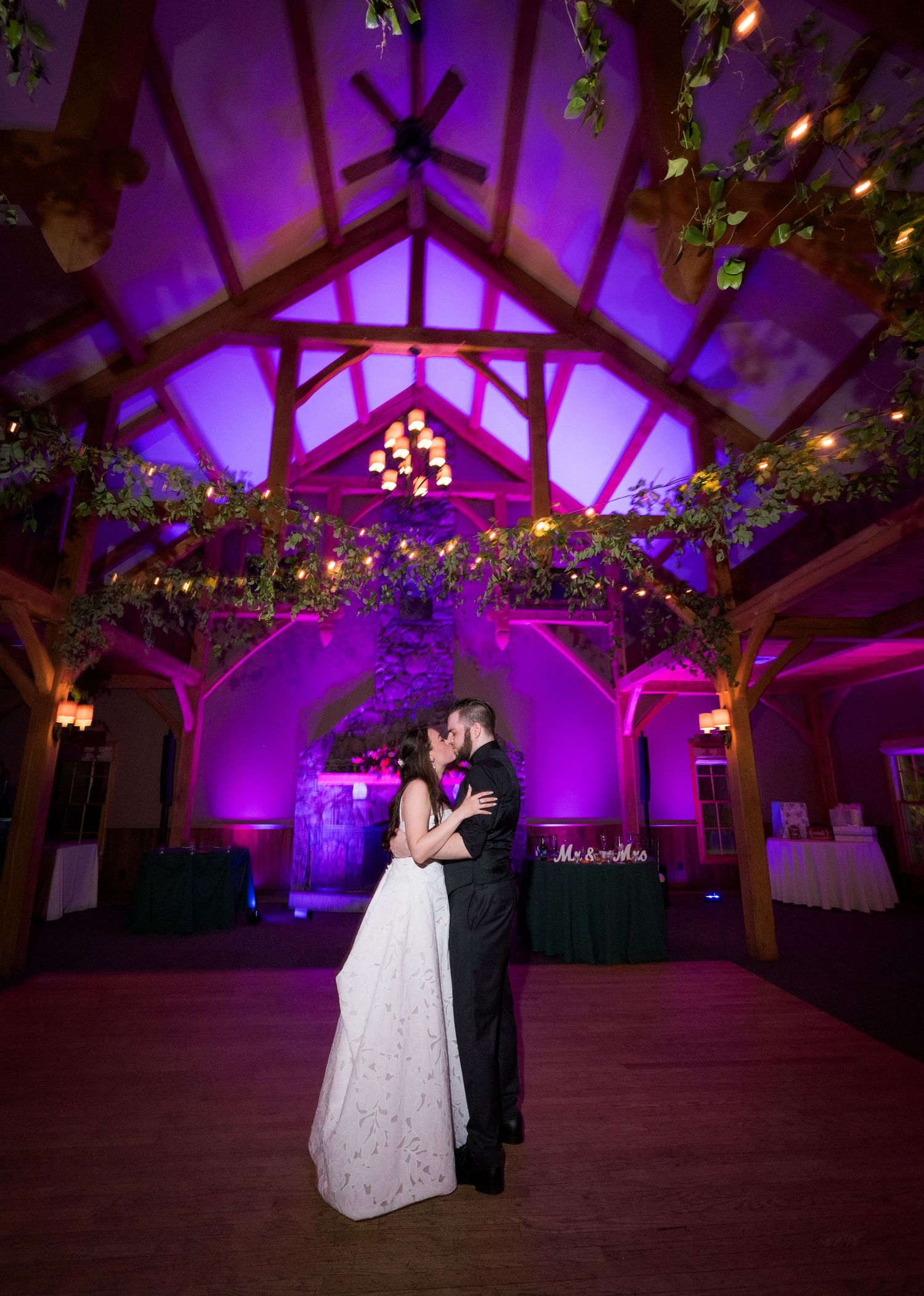 Couple shares first dance in elegant wedding venue with purple uplighting and rustic wooden beams adorned with greenery.