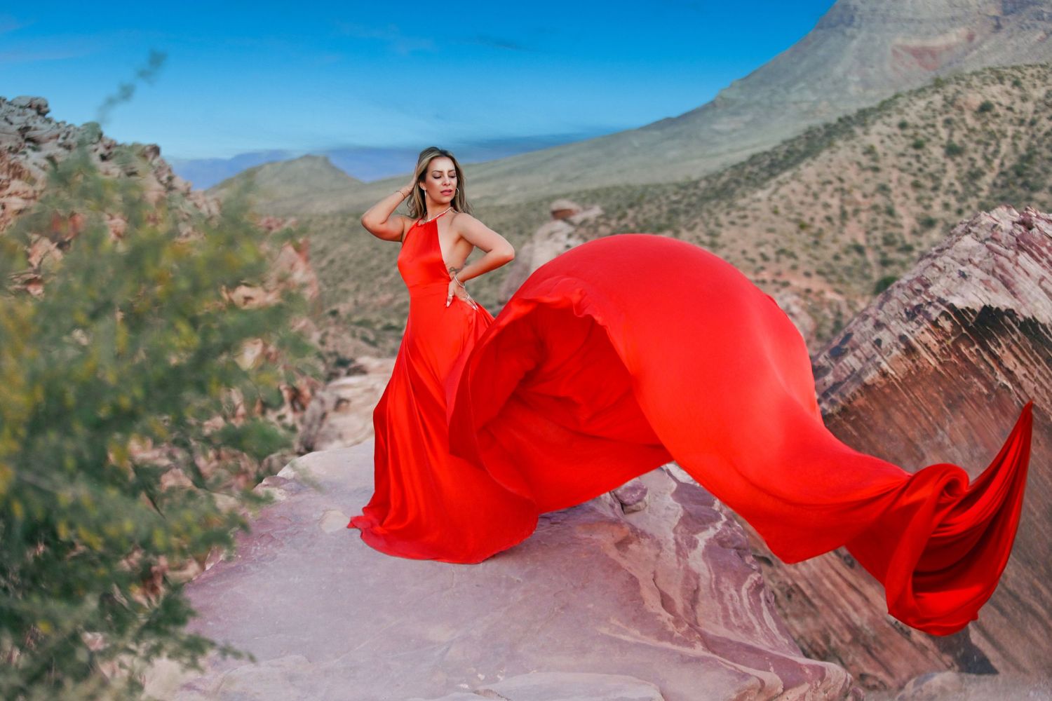 Woman in flowing red gown with dramatic fabric train posing in scenic desert mountain landscape.