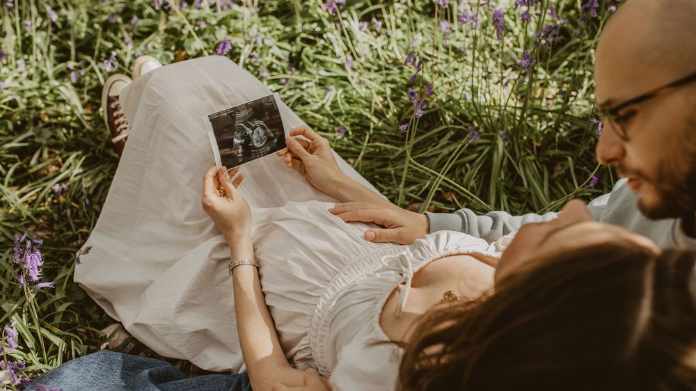 Pregnant couple lying in lavender field holding ultrasound photo, sharing tender moment outdoors.