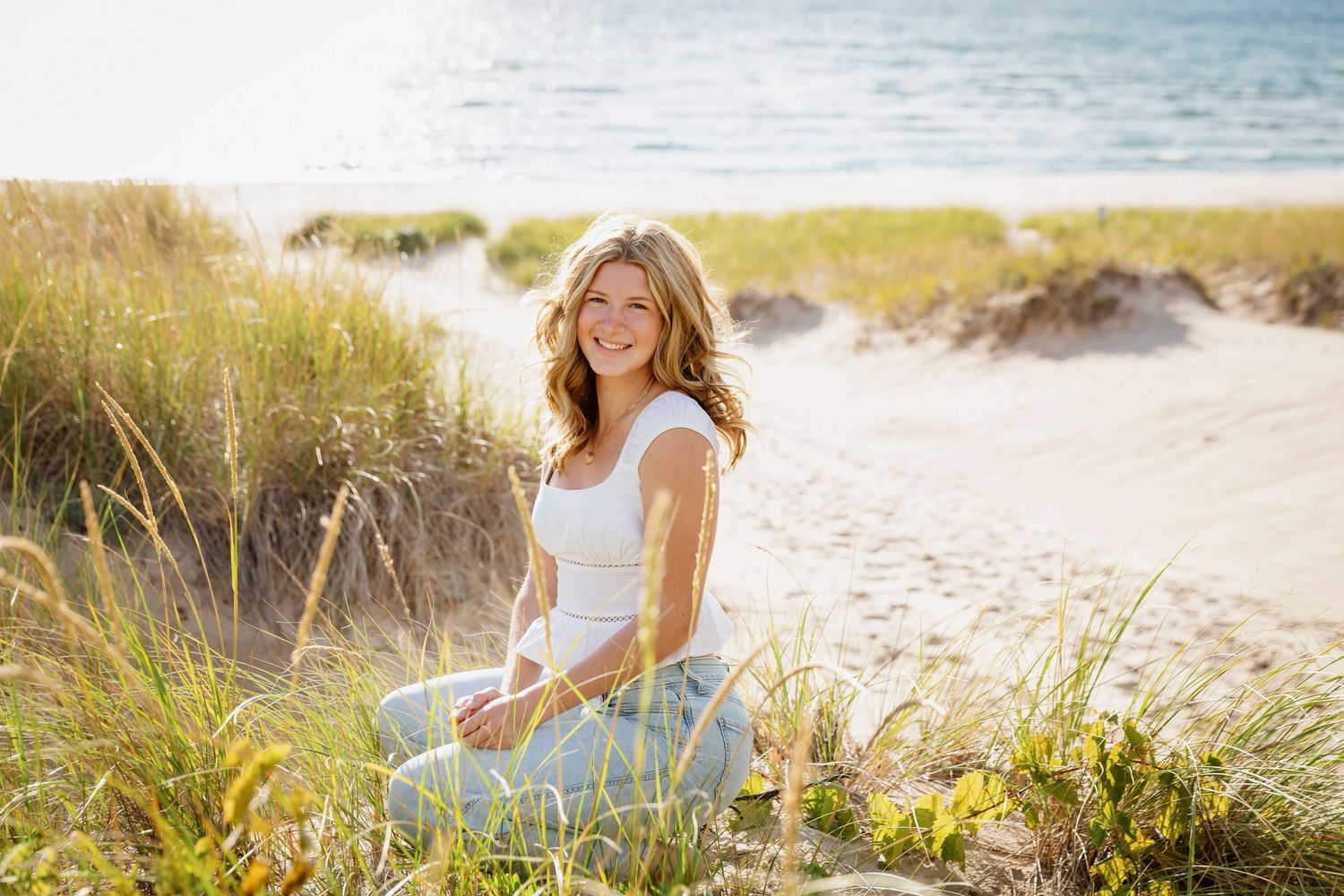 Smiling woman in white top and jeans sitting on sandy beach dunes with ocean and tall grass in background.