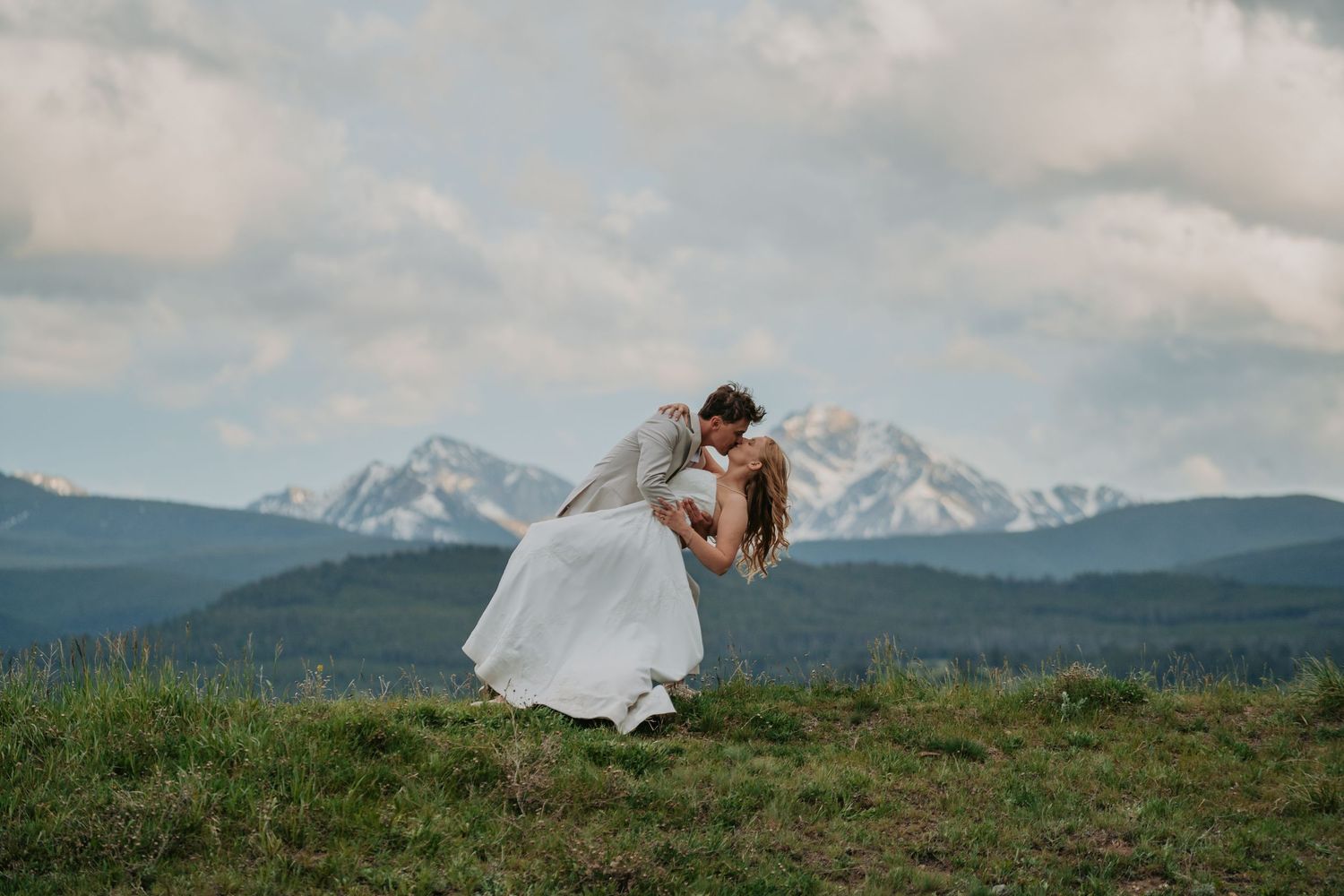 Bride and groom share a romantic dip kiss on a mountain meadow with snow-capped peaks and cloudy sky backdrop.