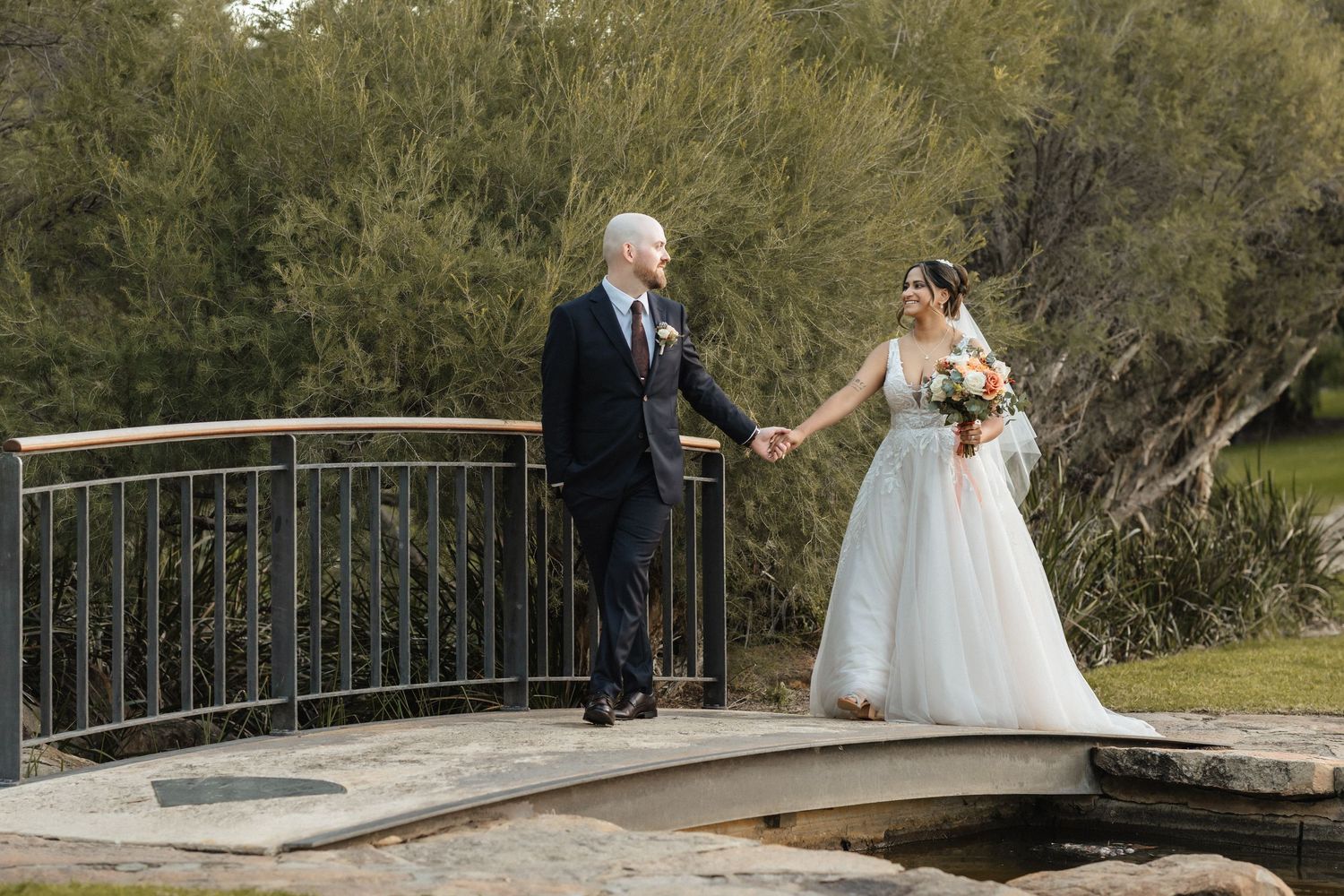 Bride and groom hold hands on a stone bridge surrounded by lush greenery on their wedding day.