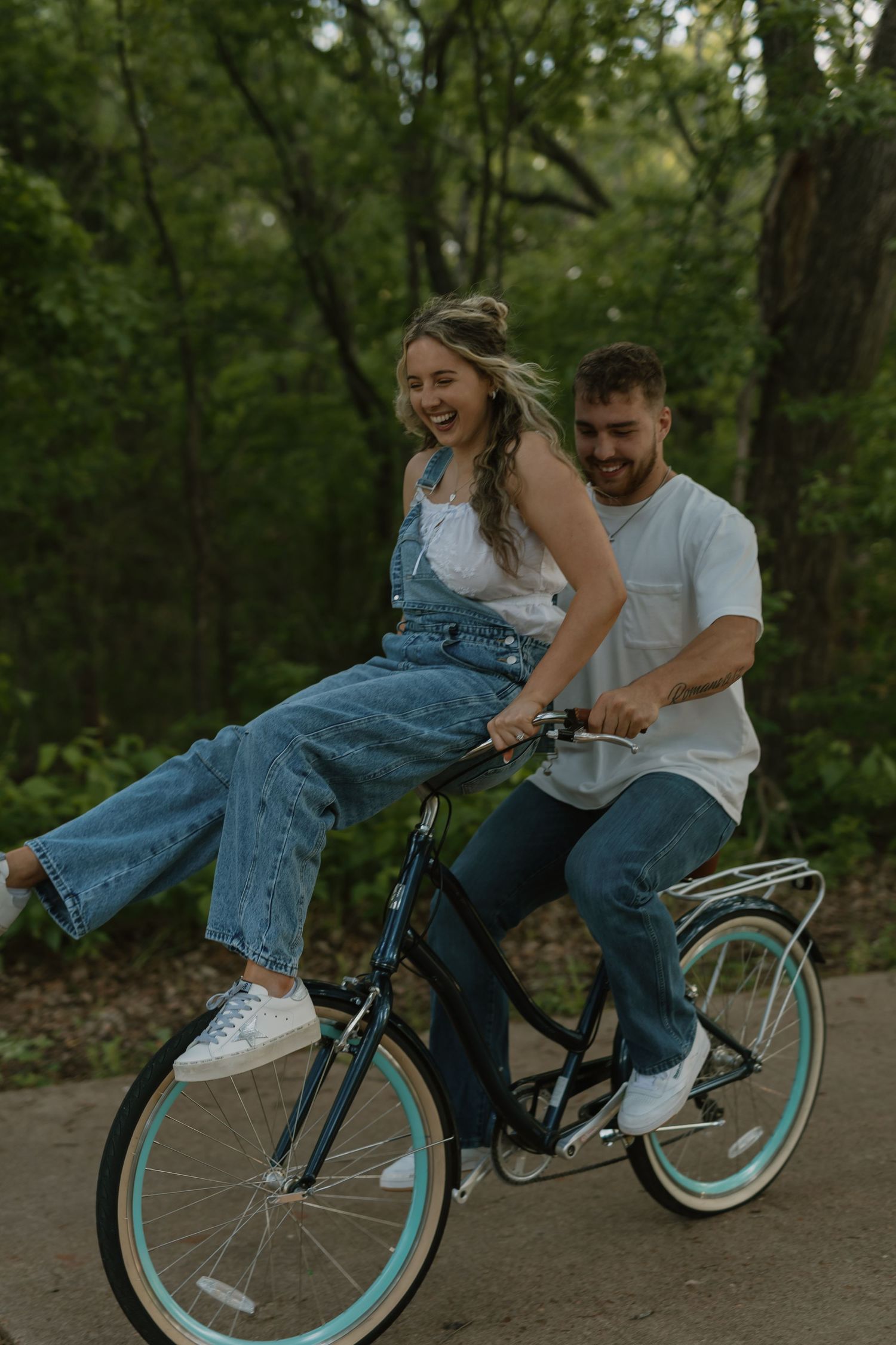 A laughing couple rides a bicycle together through a lush green forest park, woman in denim overalls sitting on handlebars.