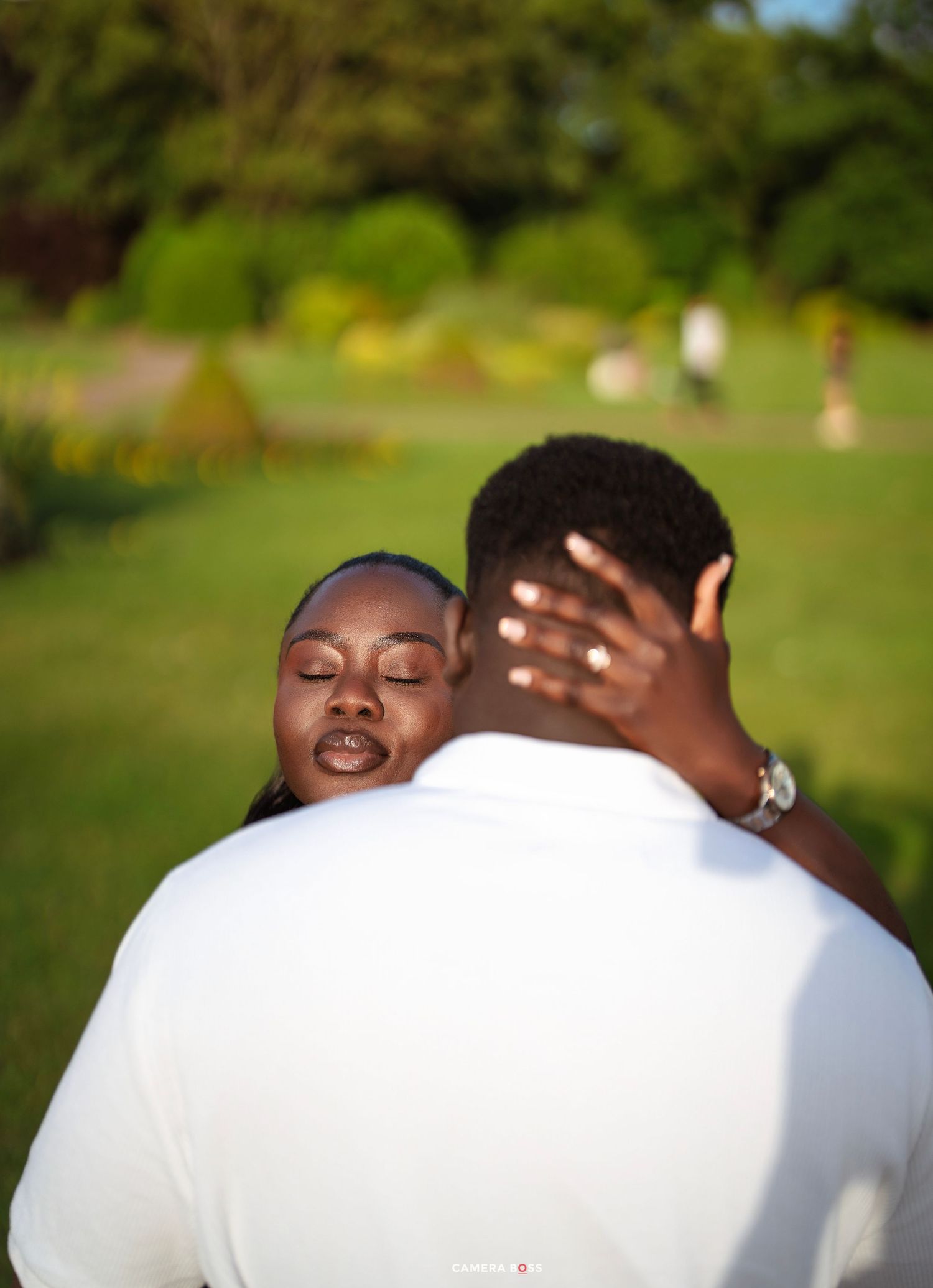 A couple embracing in a lush green park, woman with closed eyes resting her head on man's shoulder.